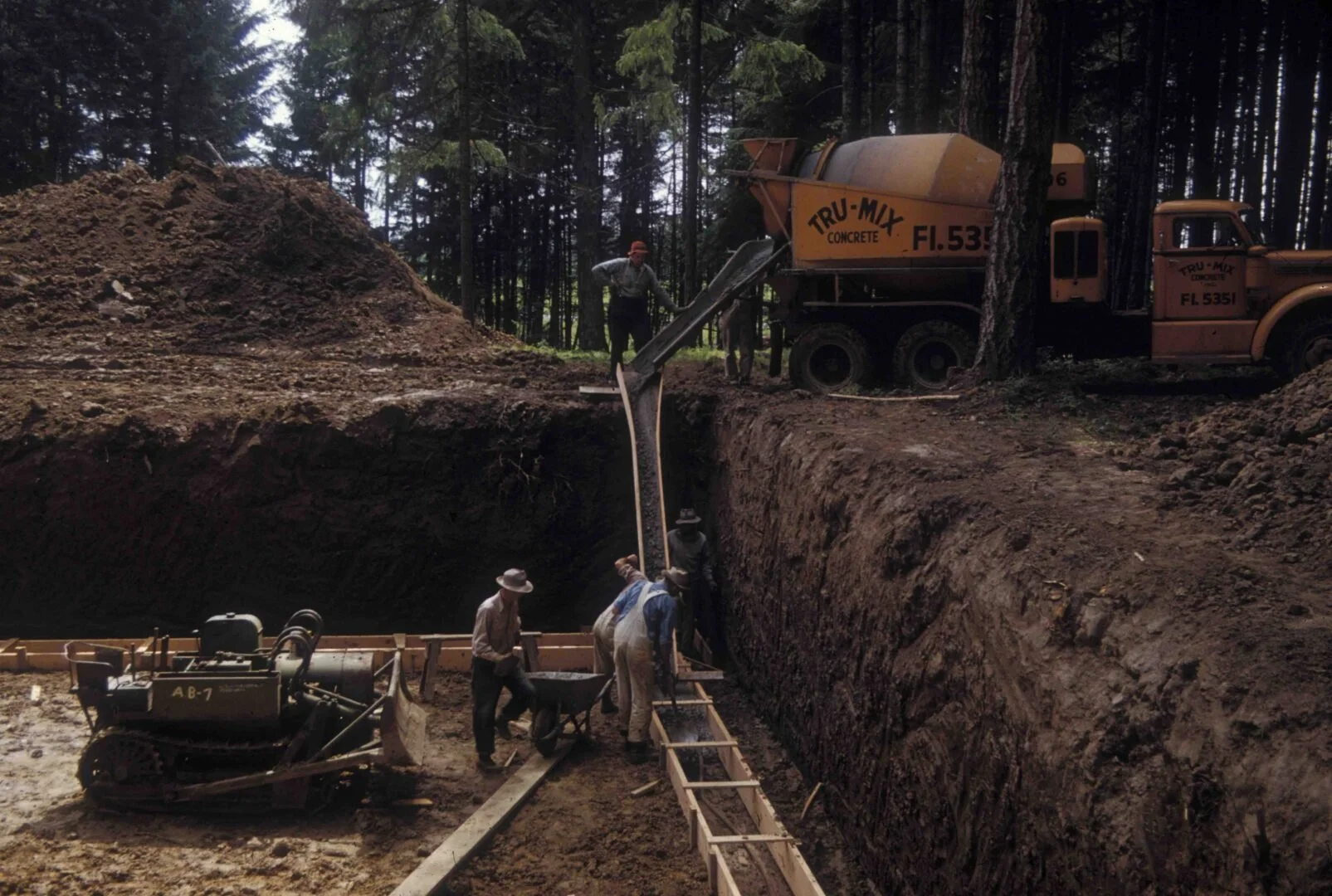 Two cement mixer trucks are parked with their pouring chutes extended over the trench. Concrete is being poured into the trench, likely to create the foundation footings or walls for the structure.