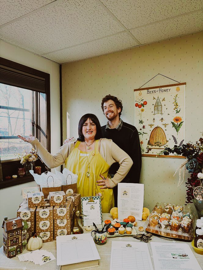 Two people standing behind a table at a honey-themed event, with honey products and baked goods displayed, and a bees and honey poster on the wall.