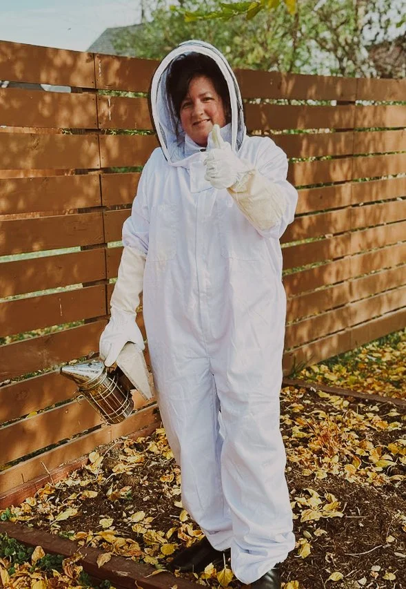 Person in a white beekeeping suit holding a honey extractor, standing outdoors near a wooden fence, with fallen leaves on the ground and trees in the background.