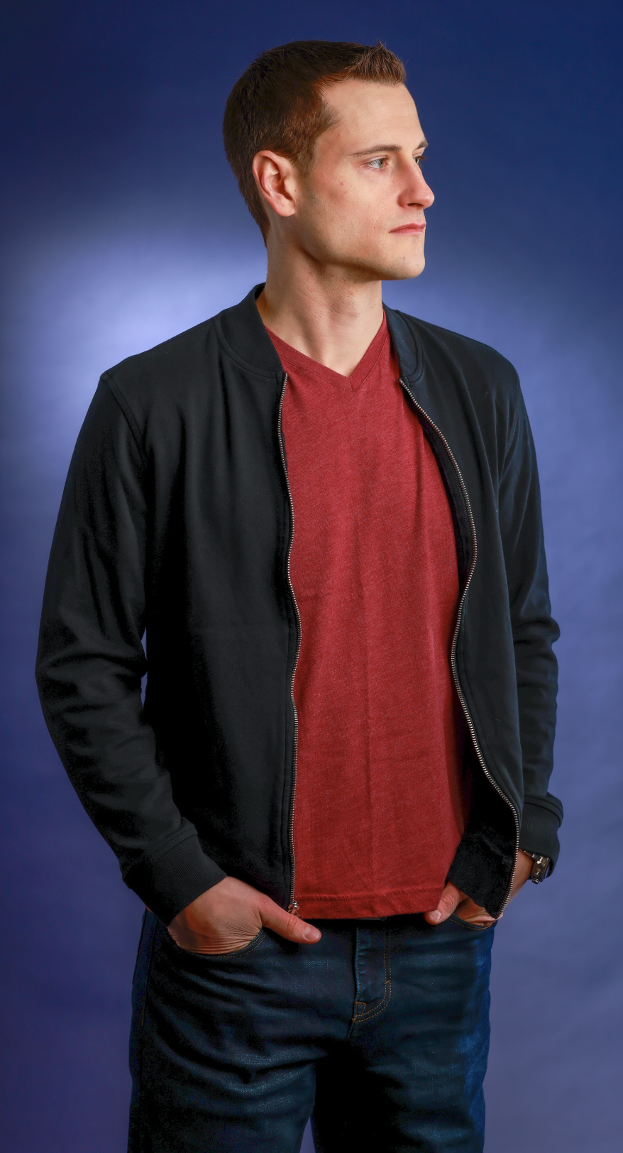 A man who is a music producer and mix engineer with short brown hair wearing a black jacket over a red t-shirt, looking to the right, standing against a blue gradient background.