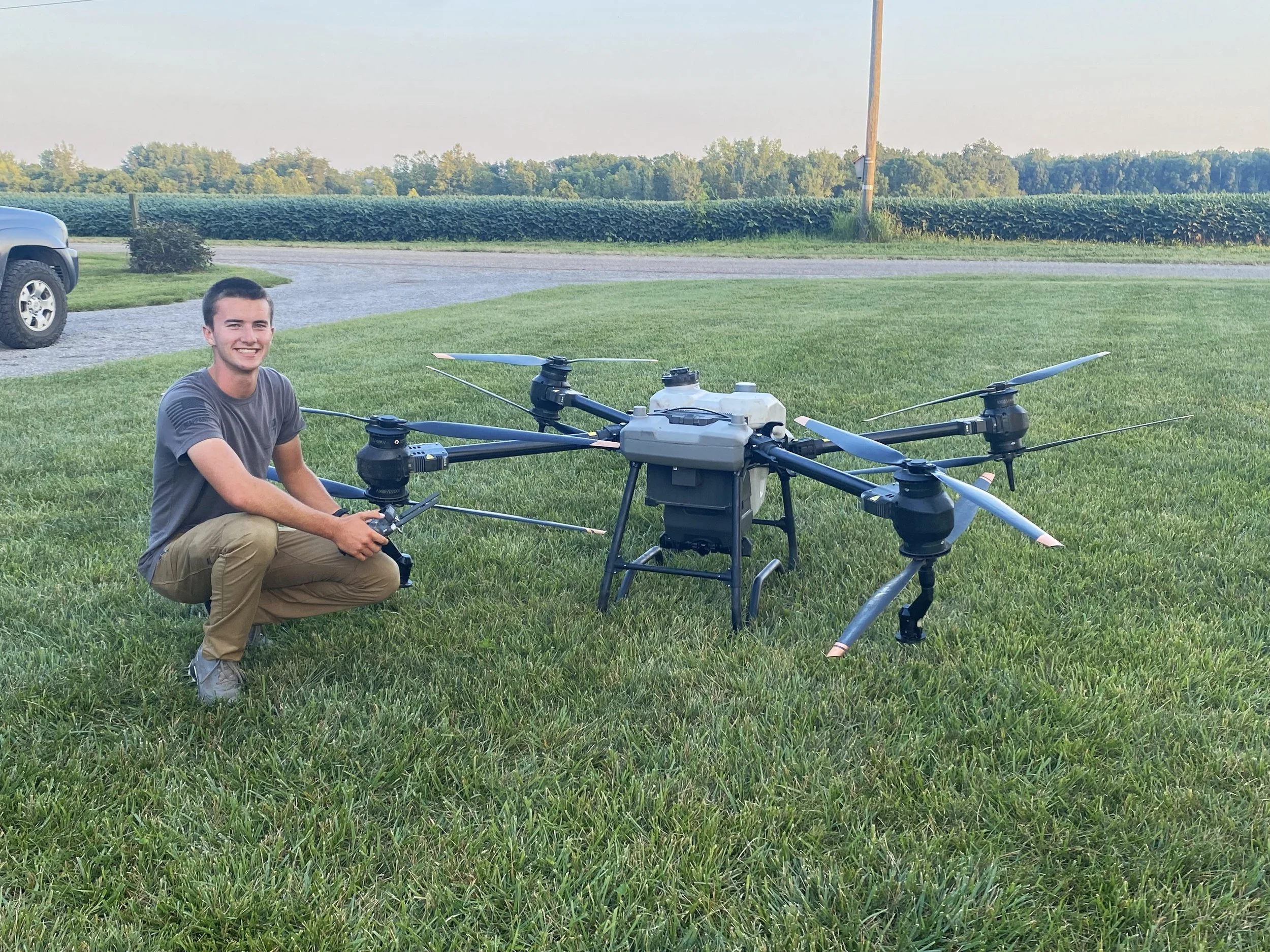 Young man crouching next to a large multirotor drone on a grassy field, with a gravel driveway, a vehicle, and farmland in the background.