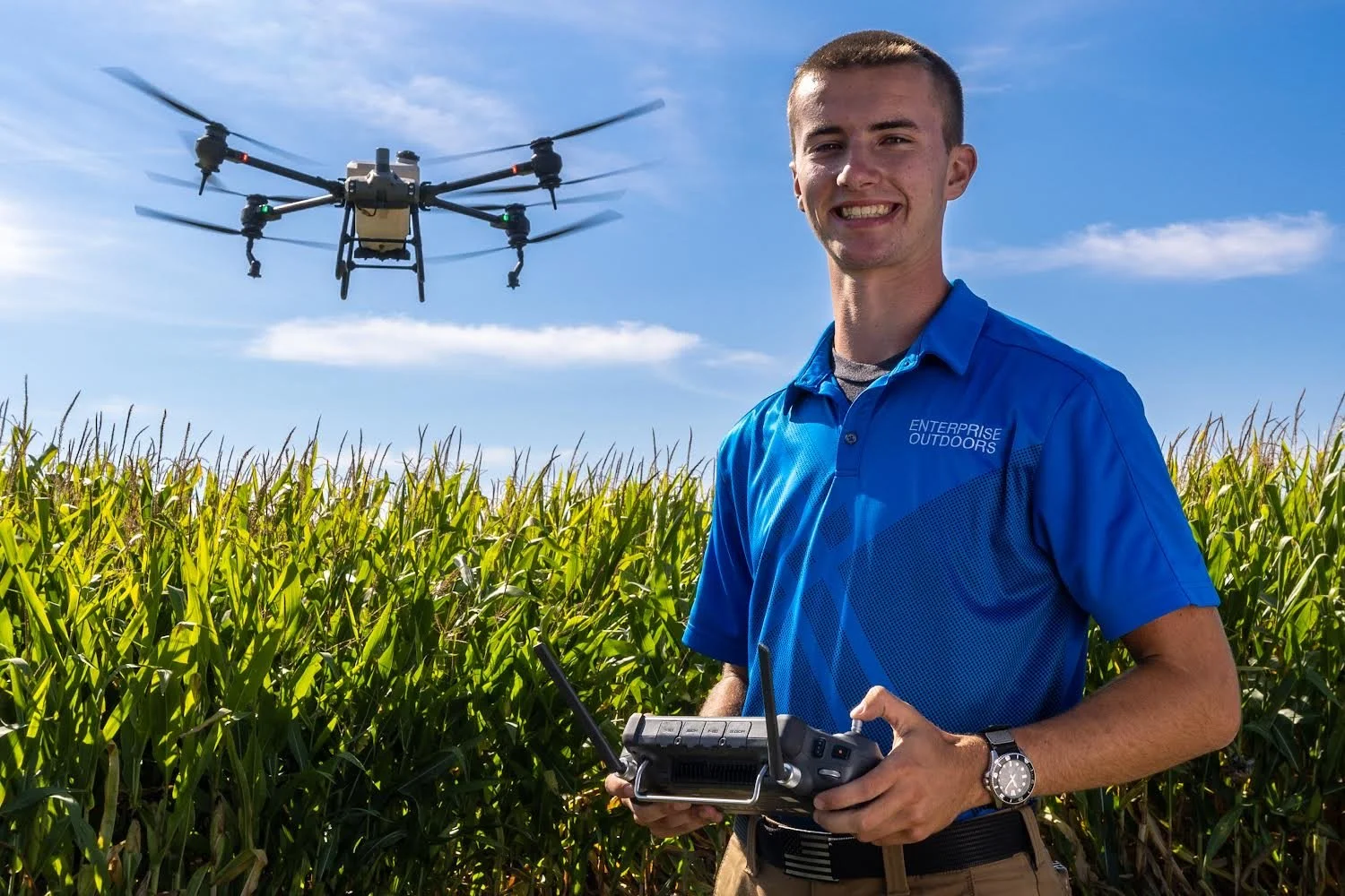 A smiling man in a blue shirt operating a drone with a remote controller in a cornfield on a sunny day.