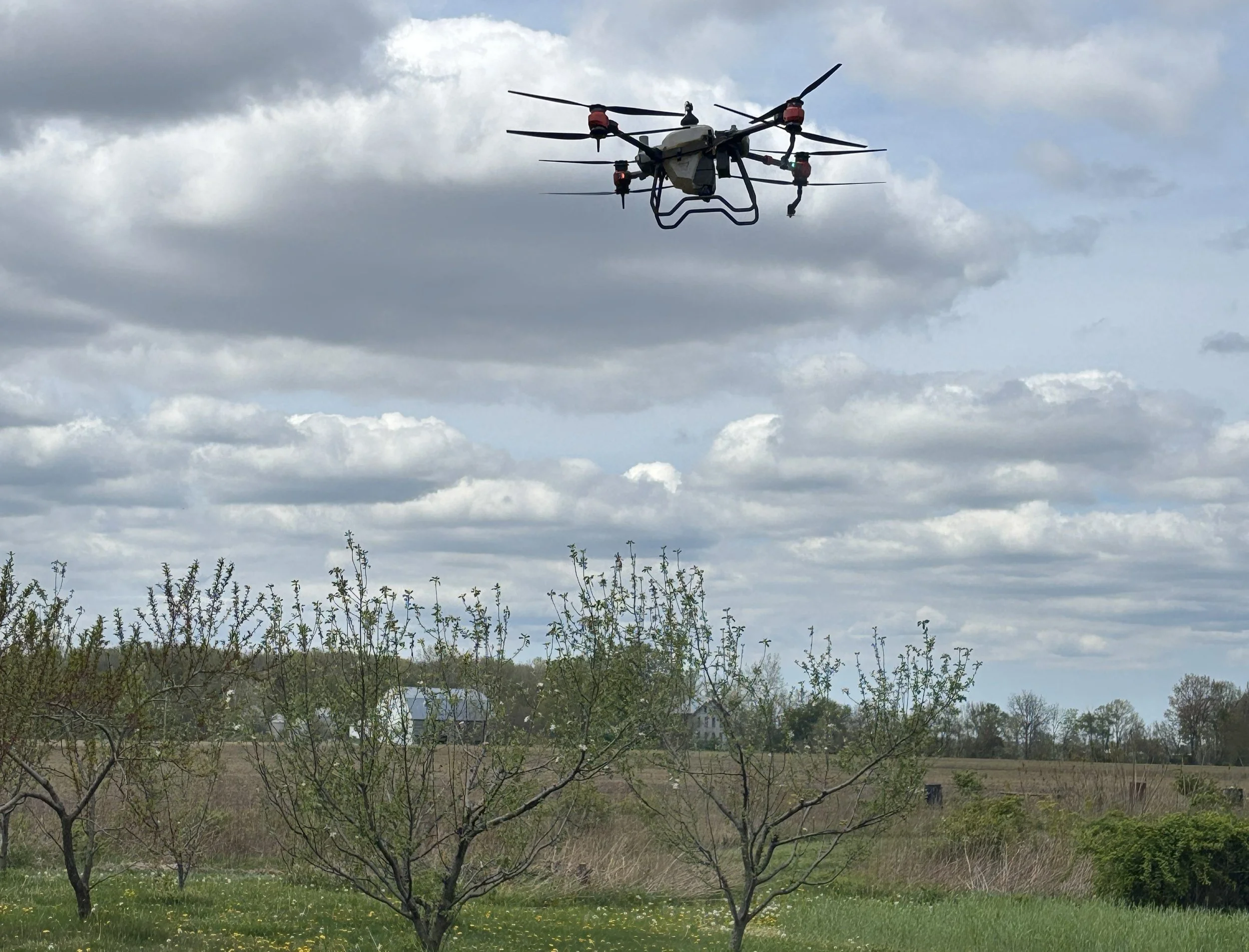 A drone flying in the sky over a rural landscape with trees and houses, under a cloudy sky.