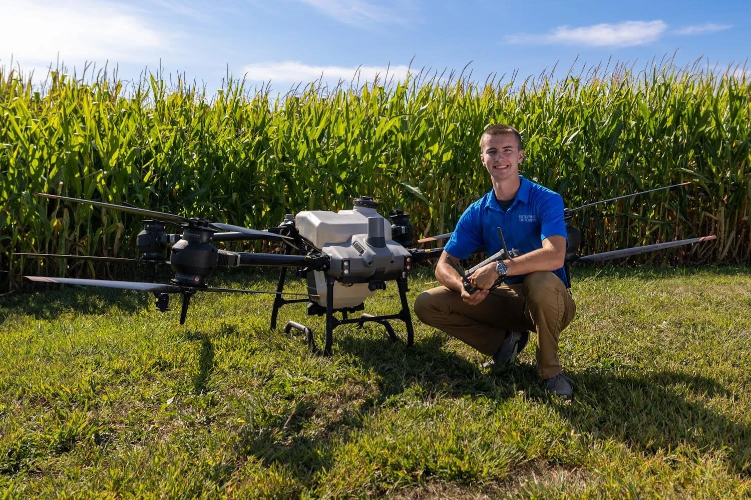 A young man in a blue shirt kneeling next to a large drone with a white container on a grassy field, with a green cornfield and blue sky in the background.
