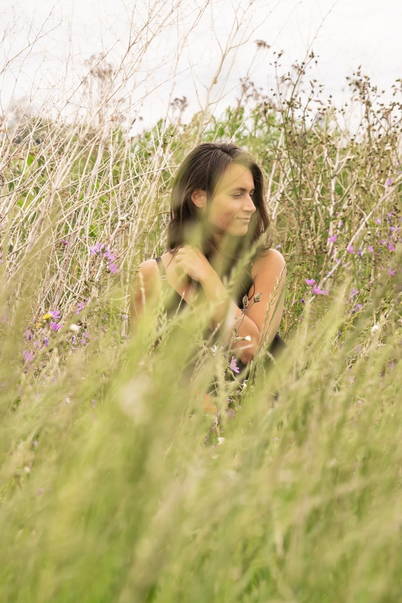 A woman with dark brown hair sitting in a grassy field with wildflowers and dried plants, looking to her left with her arms crossed.