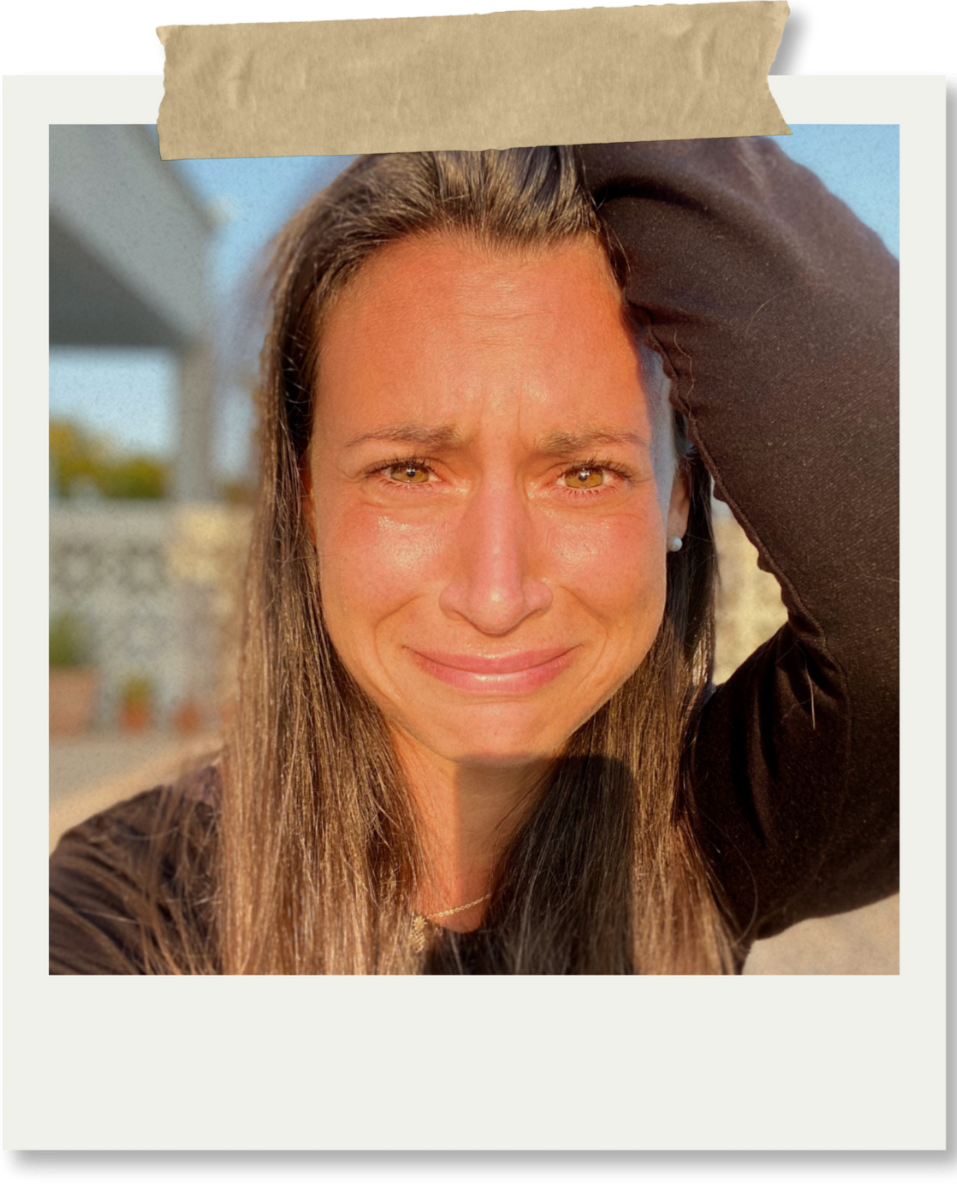 Close-up of a woman with long brown hair and brown eyes, smiling with tears on her face, holding her hand to her forehead, outdoors in sunlight.