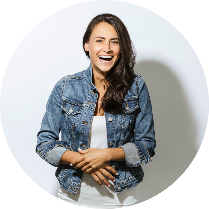 A woman laughing and smiling, wearing a denim jacket over a white shirt, with her hands clasped in front of her.
