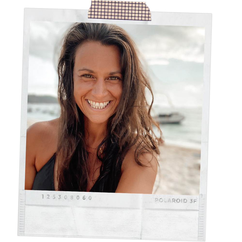 A woman with long brown hair smiling at the camera, at the beach with an ocean and a boat in the background.