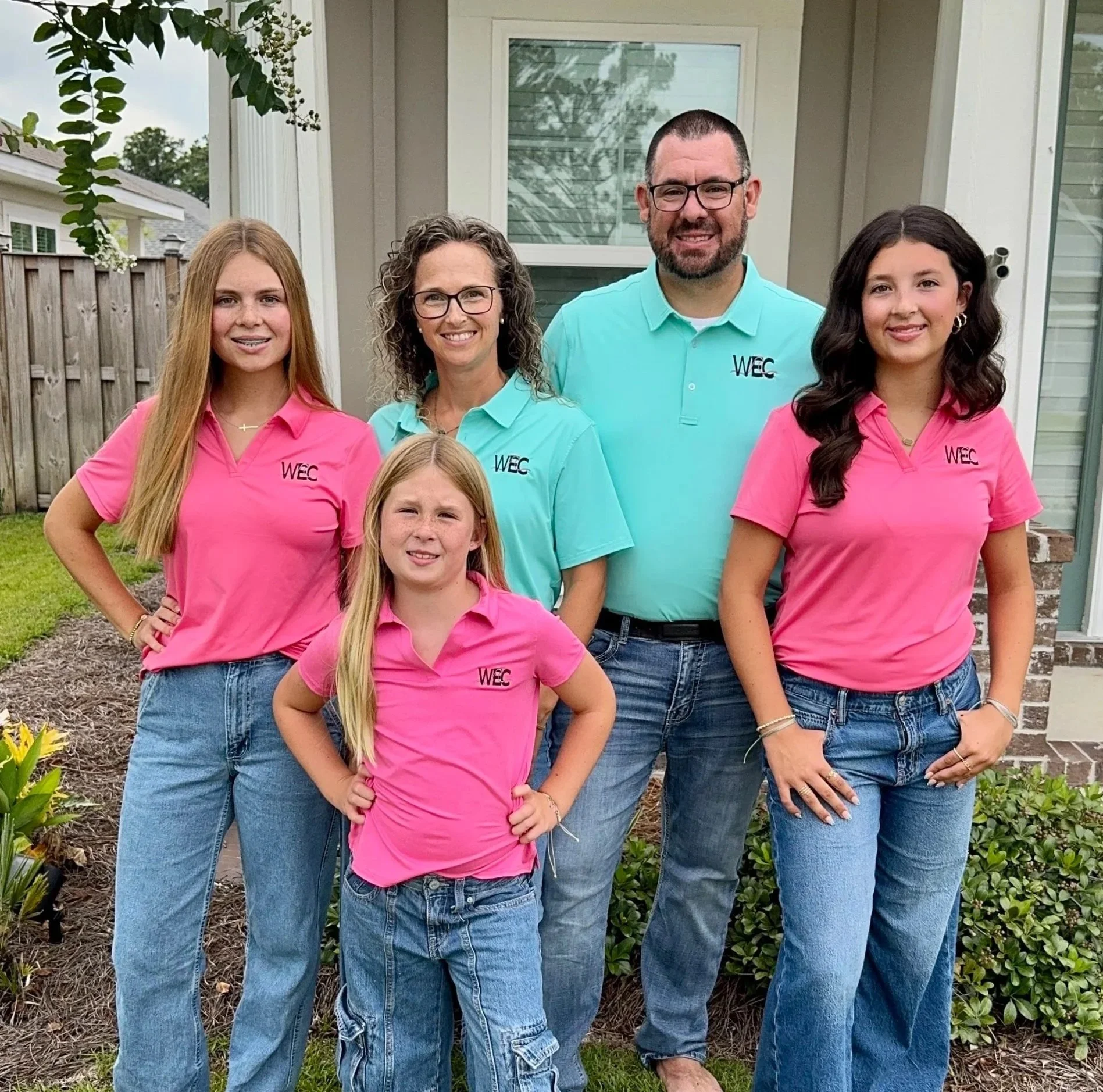 Group photo of five people standing outside in front of a house with a window and wooden fence. All are wearing pink or teal polo shirts with the 'WEC' logo, and they are smiling.