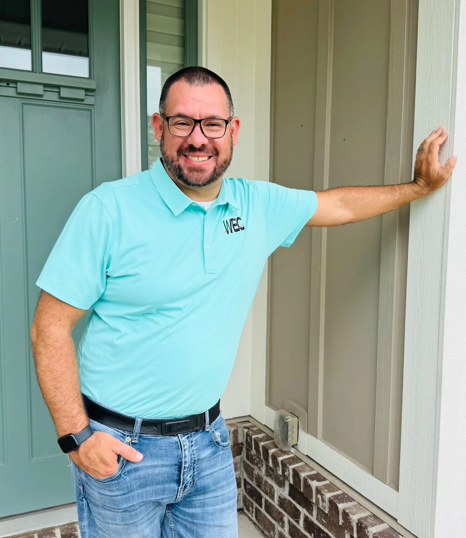 A smiling man with glasses and a beard leaning against a porch wall, wearing a light blue polo shirt with a logo, jeans, and a smartwatch.