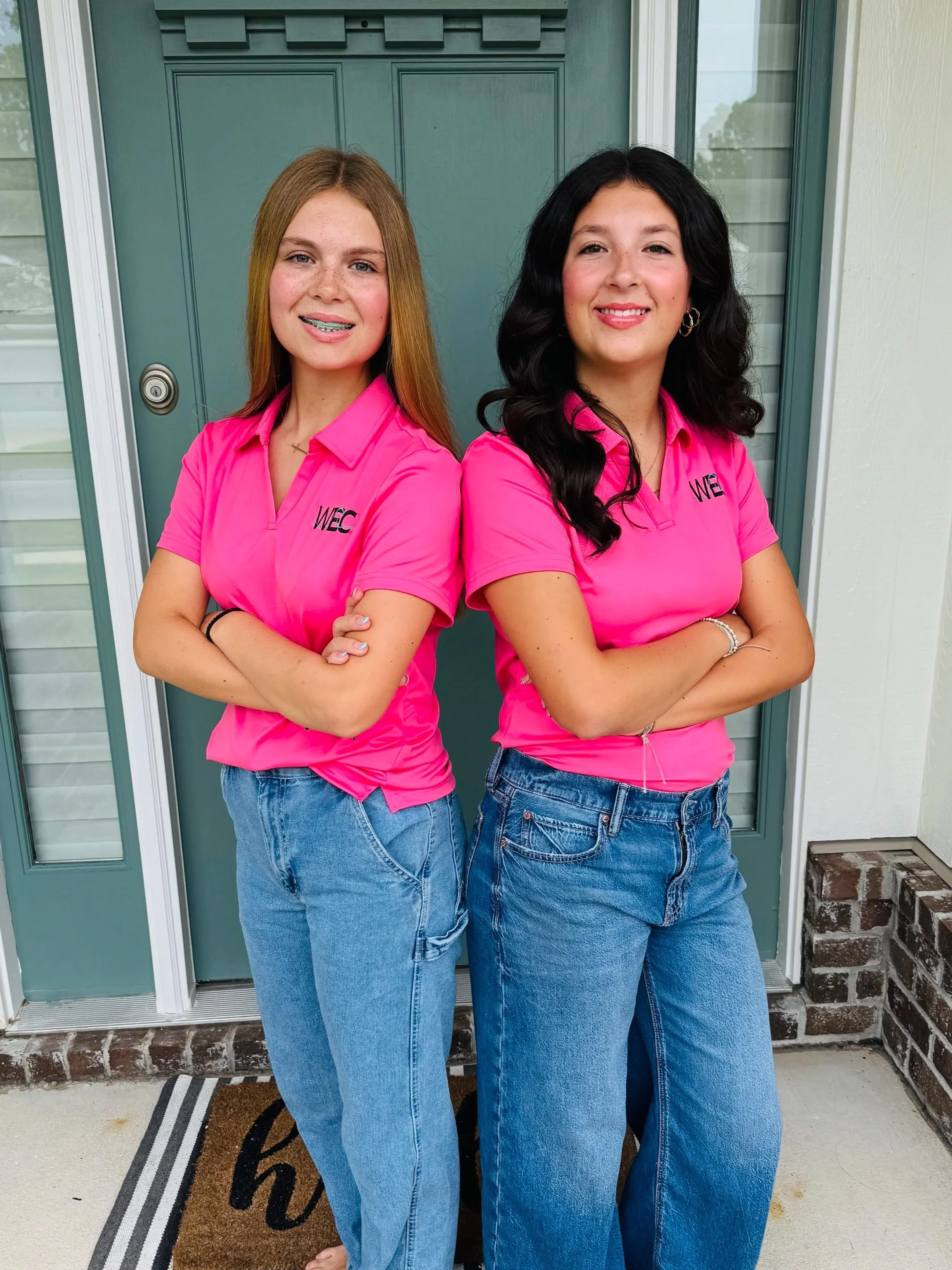 Two young women standing side by side with arms crossed, wearing matching pink shirts with 'WEC' logo, jeans, and smiling, standing in front of a green door.