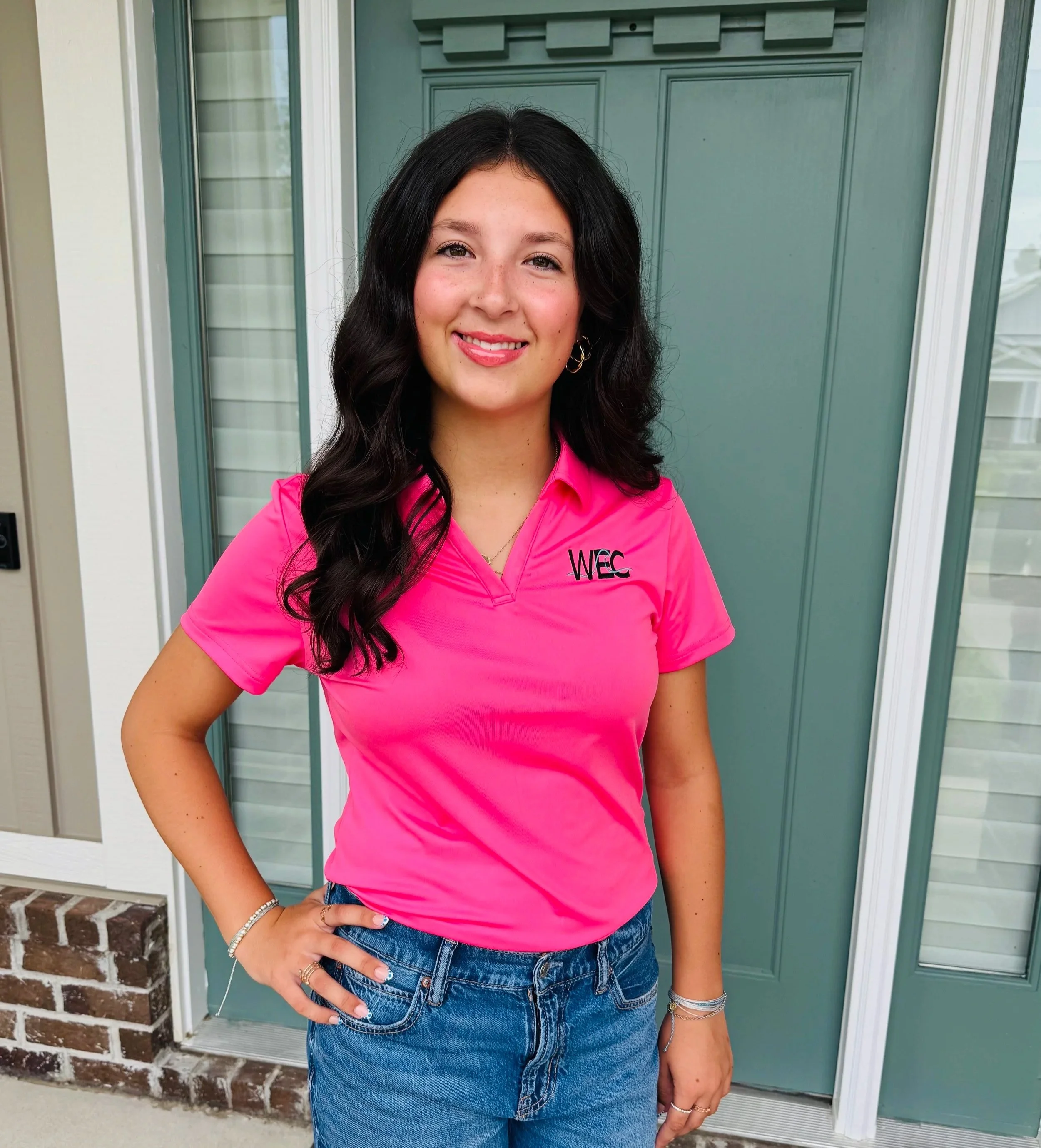 A young woman with long dark hair, wearing a bright pink polo shirt with 'WEC' embroidered on it, and blue jeans, standing outside a house with a teal door and window, smiling at the camera.