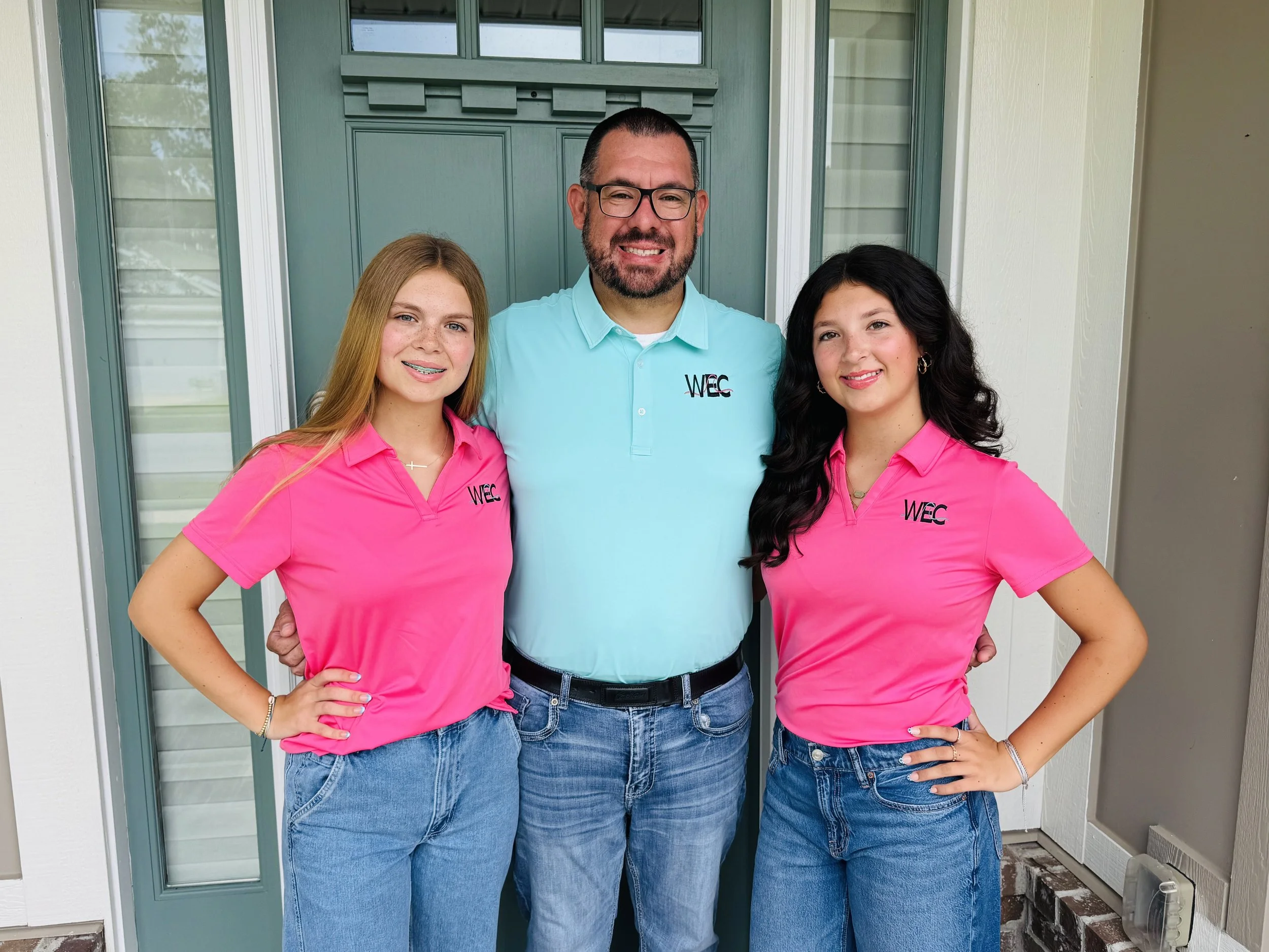 Three smiling people, two young women and one man, standing outside a house in front of a teal-colored door. The women are wearing bright pink polo shirts with the WEC logo, and the man is wearing a light blue polo shirt with the WEC logo. The women have their hands on their hips, and the man has his arms around their waists.