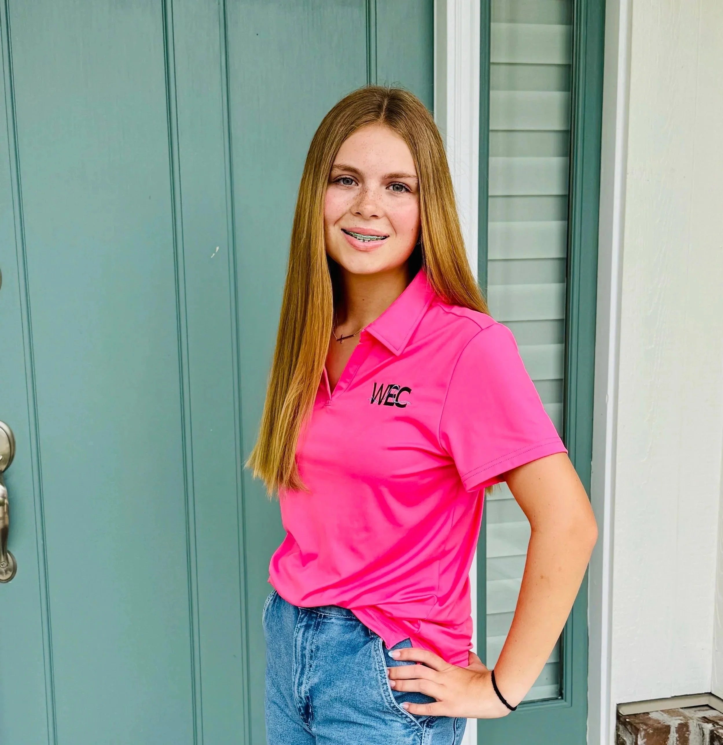 A teenage girl with long red hair, braces, and freckles standing in front of a teal door, wearing a bright pink collared shirt with 'WEC' embroidered on it and blue jeans.