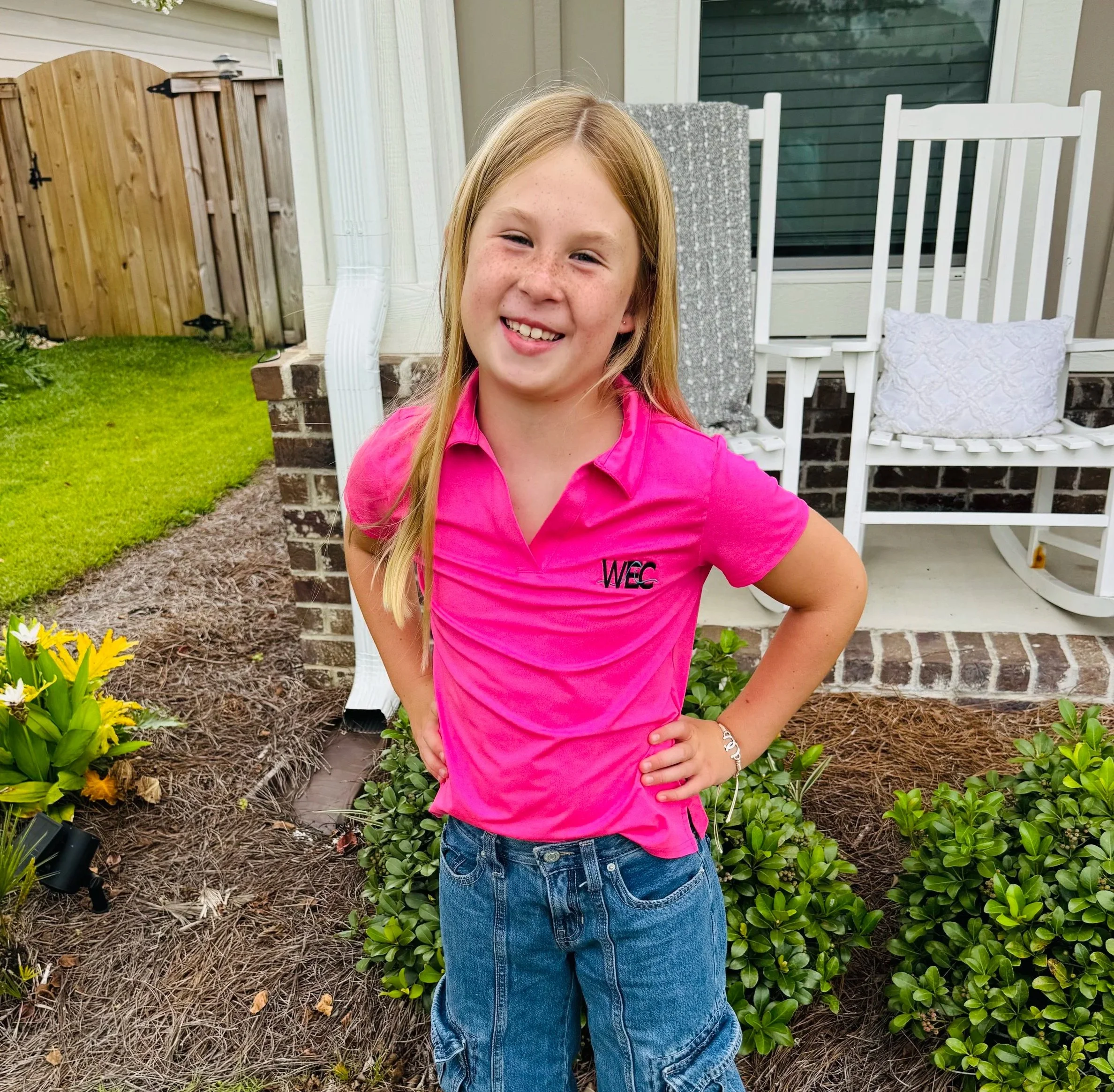 A girl with long blonde hair in a pink polo shirt standing outside on a porch with hands on hips, smiling. There are plants, grass, and a white porch chair in the background.