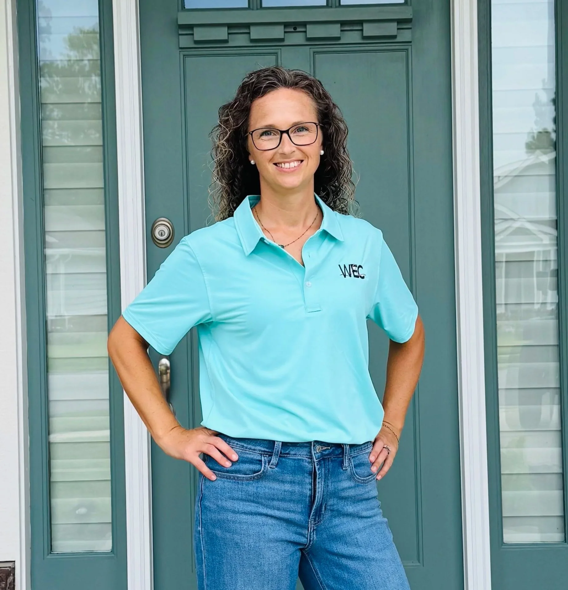 A woman with curly brown hair and glasses standing with her hand on her hip in front of a green door, wearing a turquoise polo shirt with a logo and blue jeans.