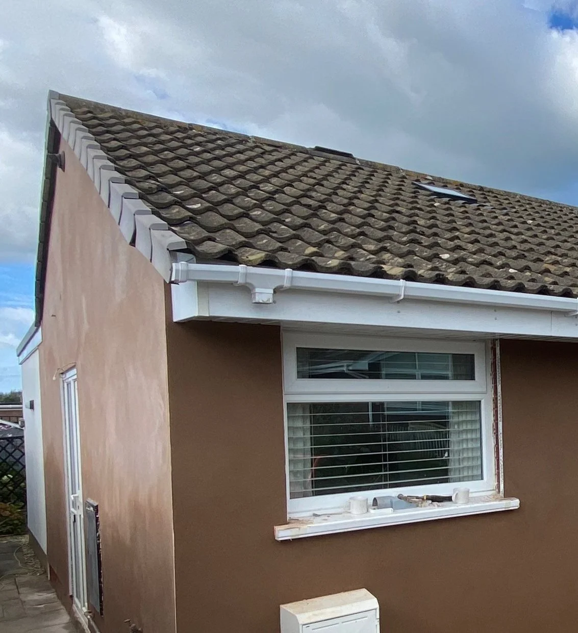 The side of a house with a brown exterior wall, a white-framed window with blinds, and a tiled roof with a small skylight. A white gutter system runs along the roof edge.