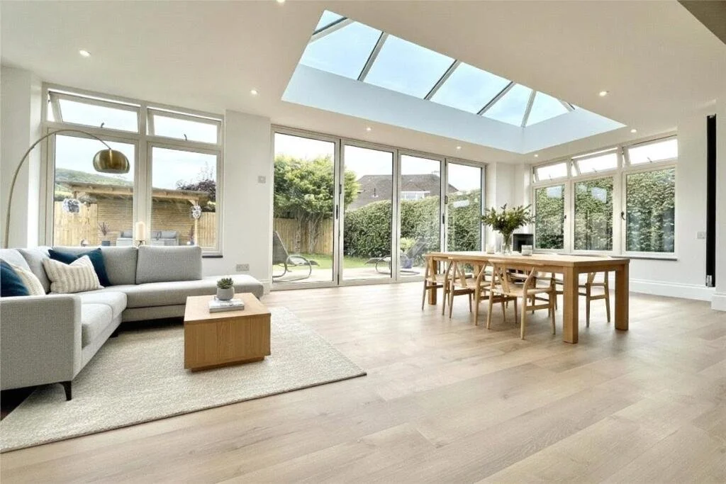 A spacious living and dining area with large windows and a skylight, featuring a white sofa, wooden coffee table, dining table with chairs, and garden view outside.