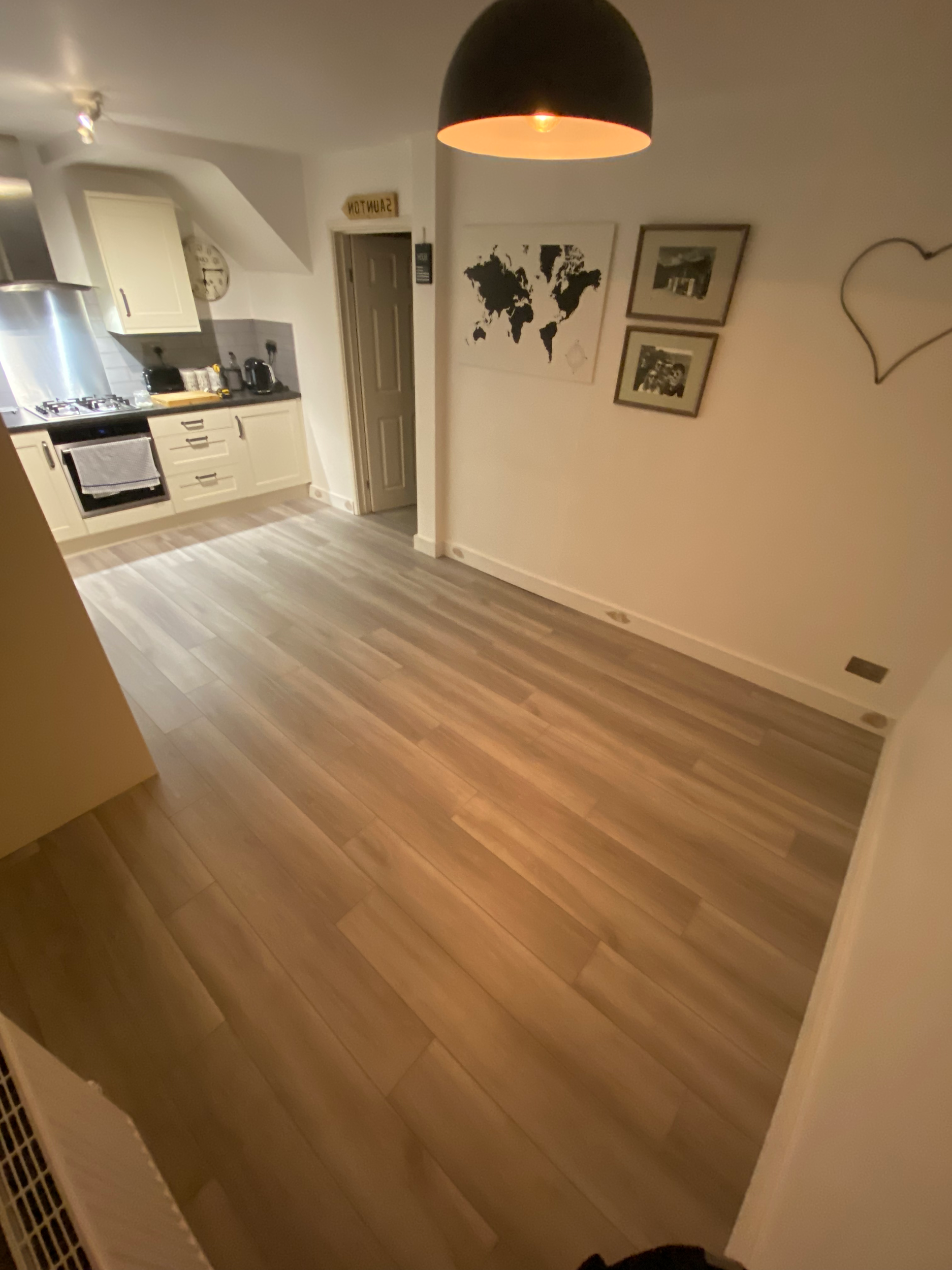 View of a kitchen with white cabinets, a stove, a sink, and countertops. The room features wooden flooring and wall decor including a world map and framed photos. Part of a dark hanging lamp and an abstract wall outline can be seen.