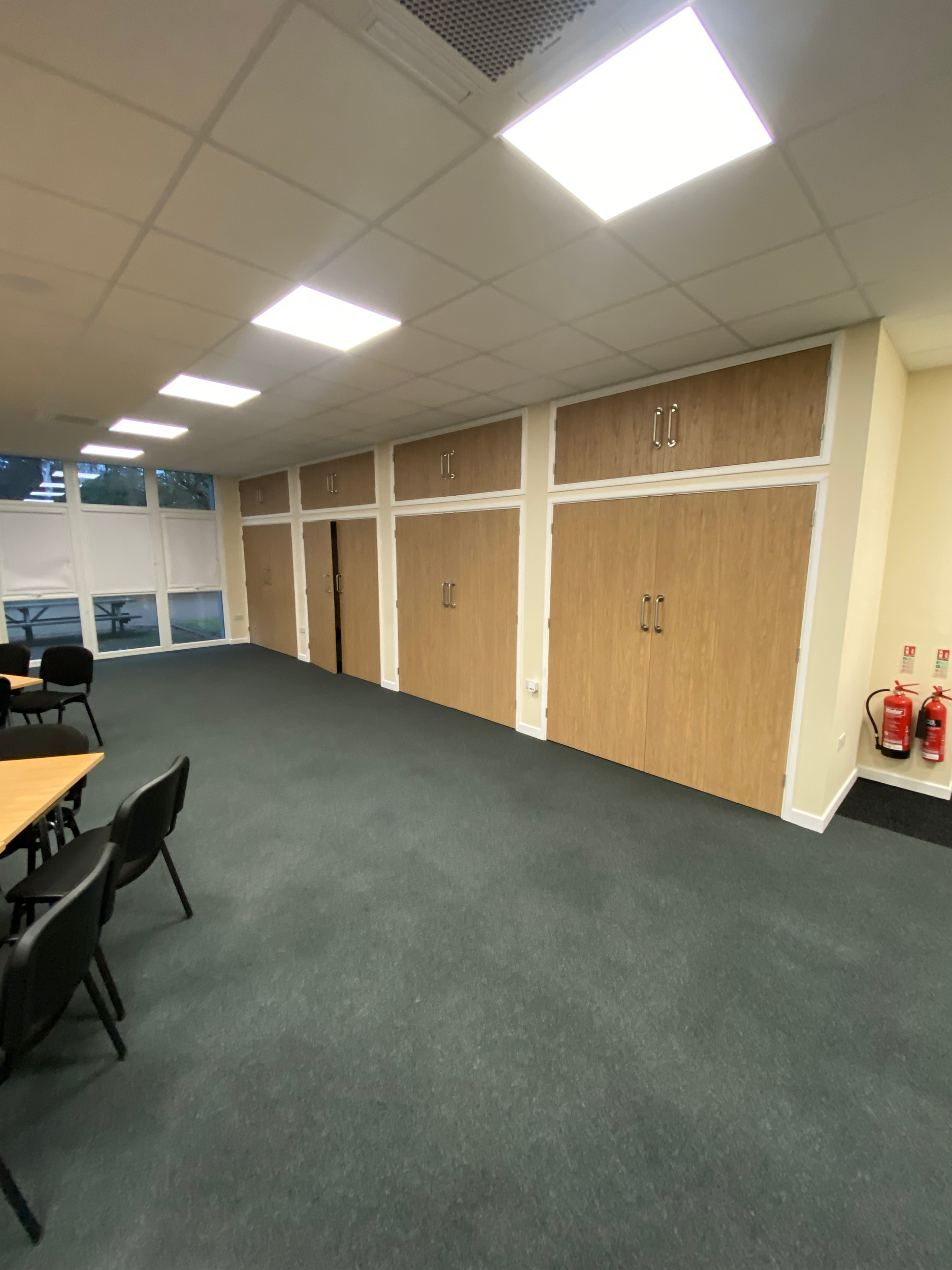 Empty office room with wooden cabinets, black chairs, a table, and fire extinguishers on the wall. Fluorescent lighting on the ceiling and windows along one wall.