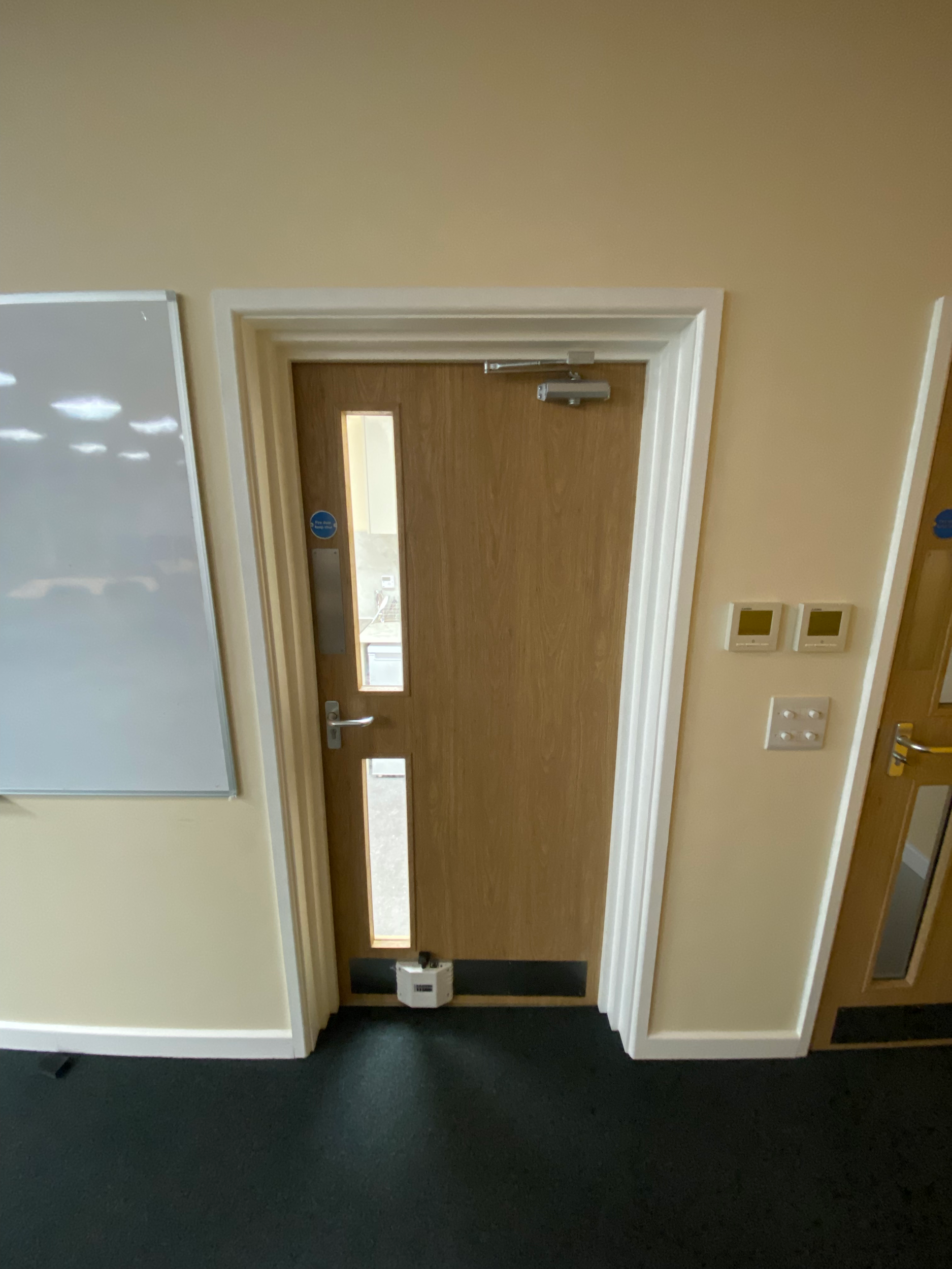 Wooden door with a vertical window, door handle, and a push bar, set in a beige wall with electrical switches and a fire alarm, adjacent to a whiteboard and a dark-colored floor.