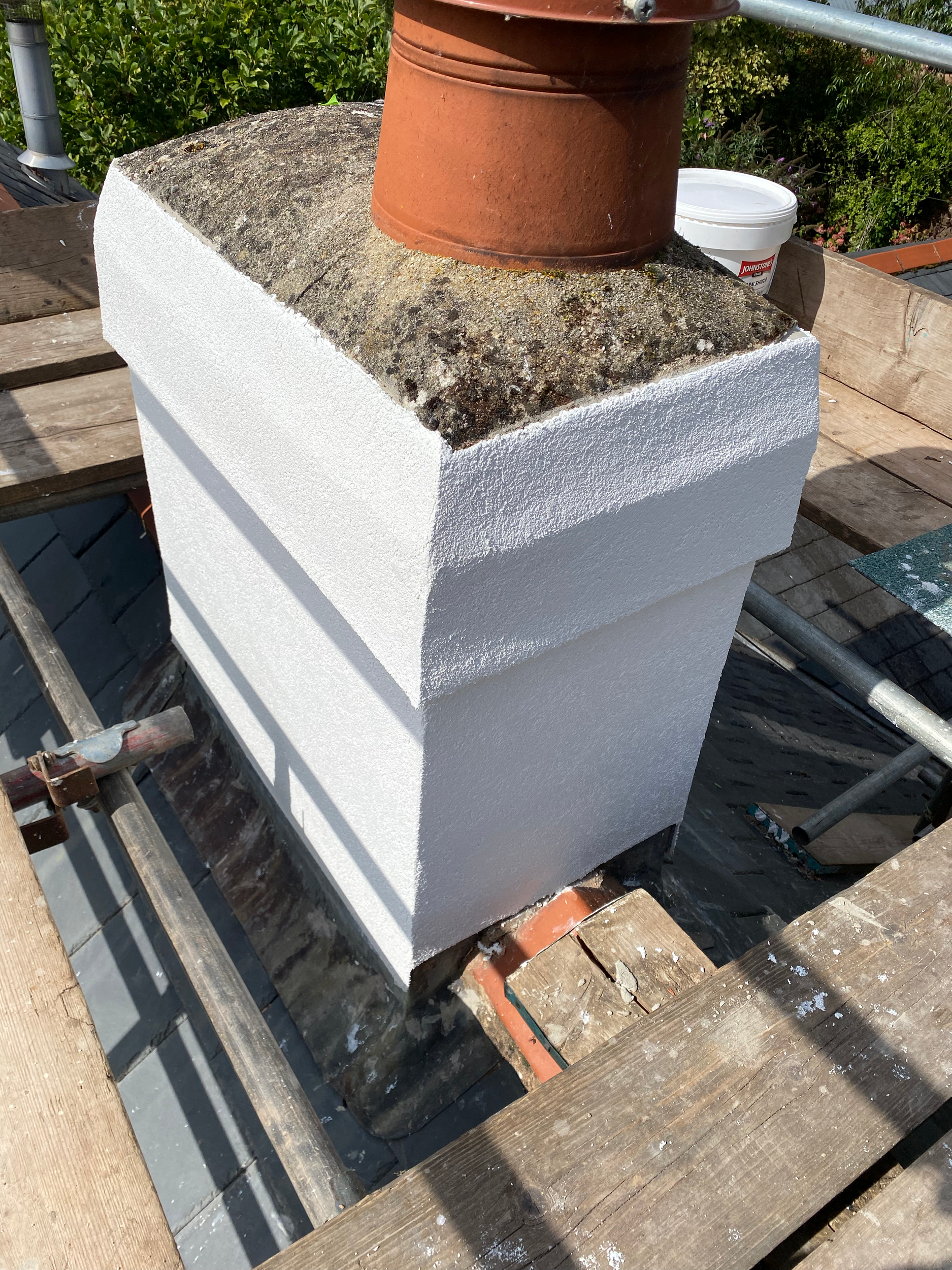 A white-painted chimney on a rooftop with a terra cotta flowerpot resting on top. The roof has a wooden surface with scaffolding around it, and other gardening items are visible in the background.