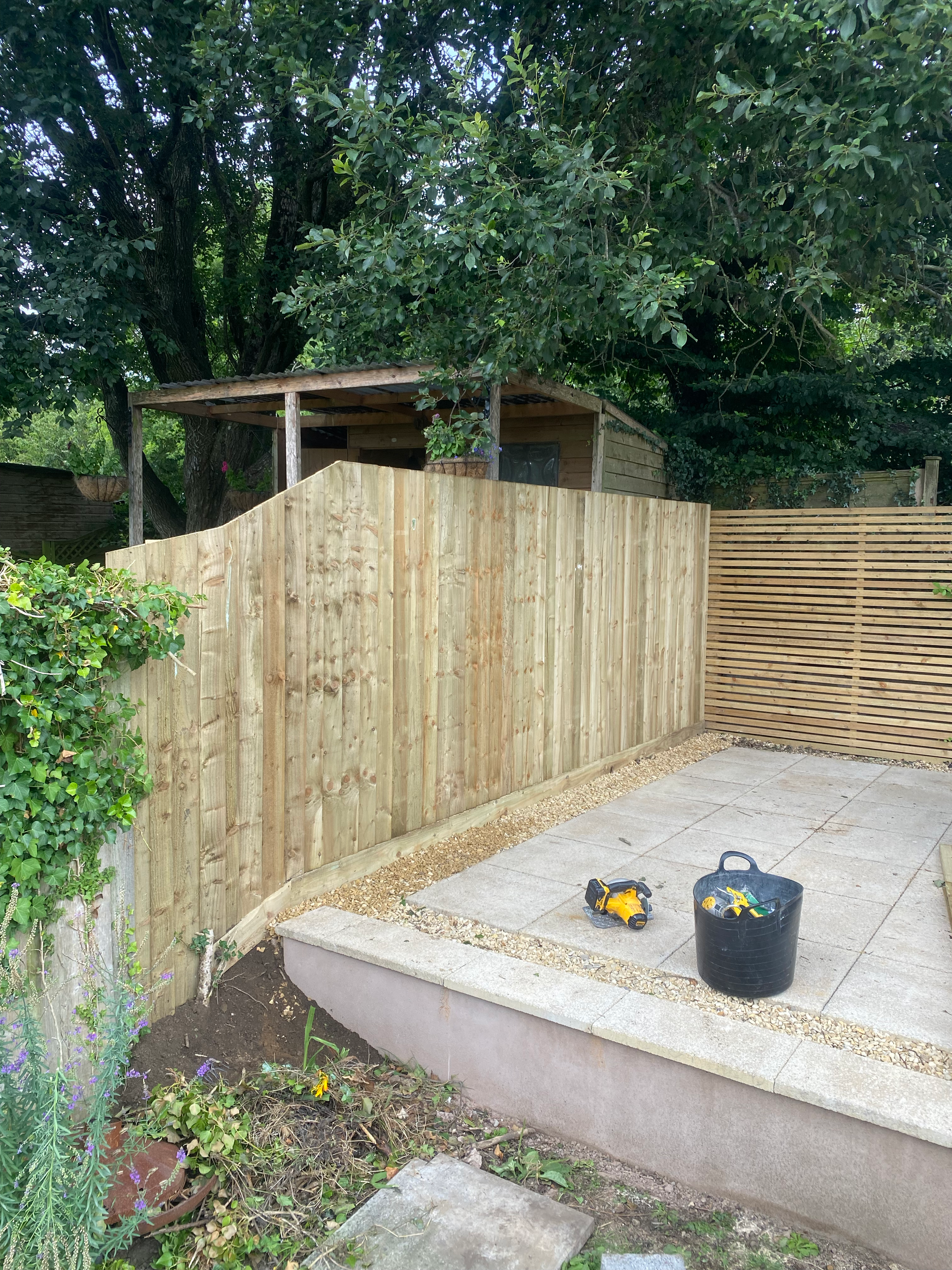 A backyard with a partially built wooden fence, a paved patio area, and gardening tools and supplies. Green trees and shrubs surround the space.