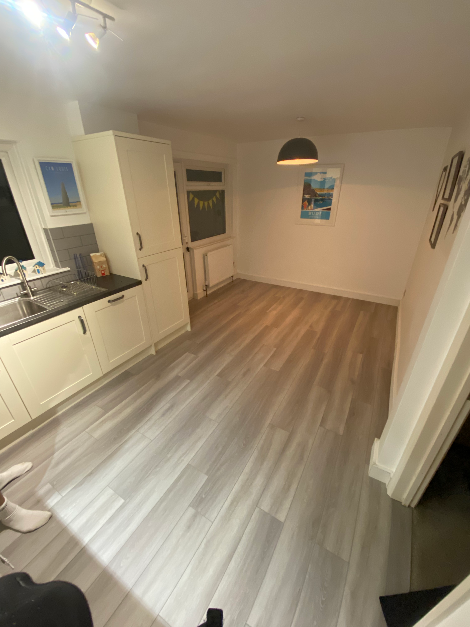A kitchen with white cabinets, gray countertops, a window with a sun design on the blinds, and light wood flooring. A poster of a lighthouse on the wall, a hanging black pendant light, and a person wearing white socks are visible.