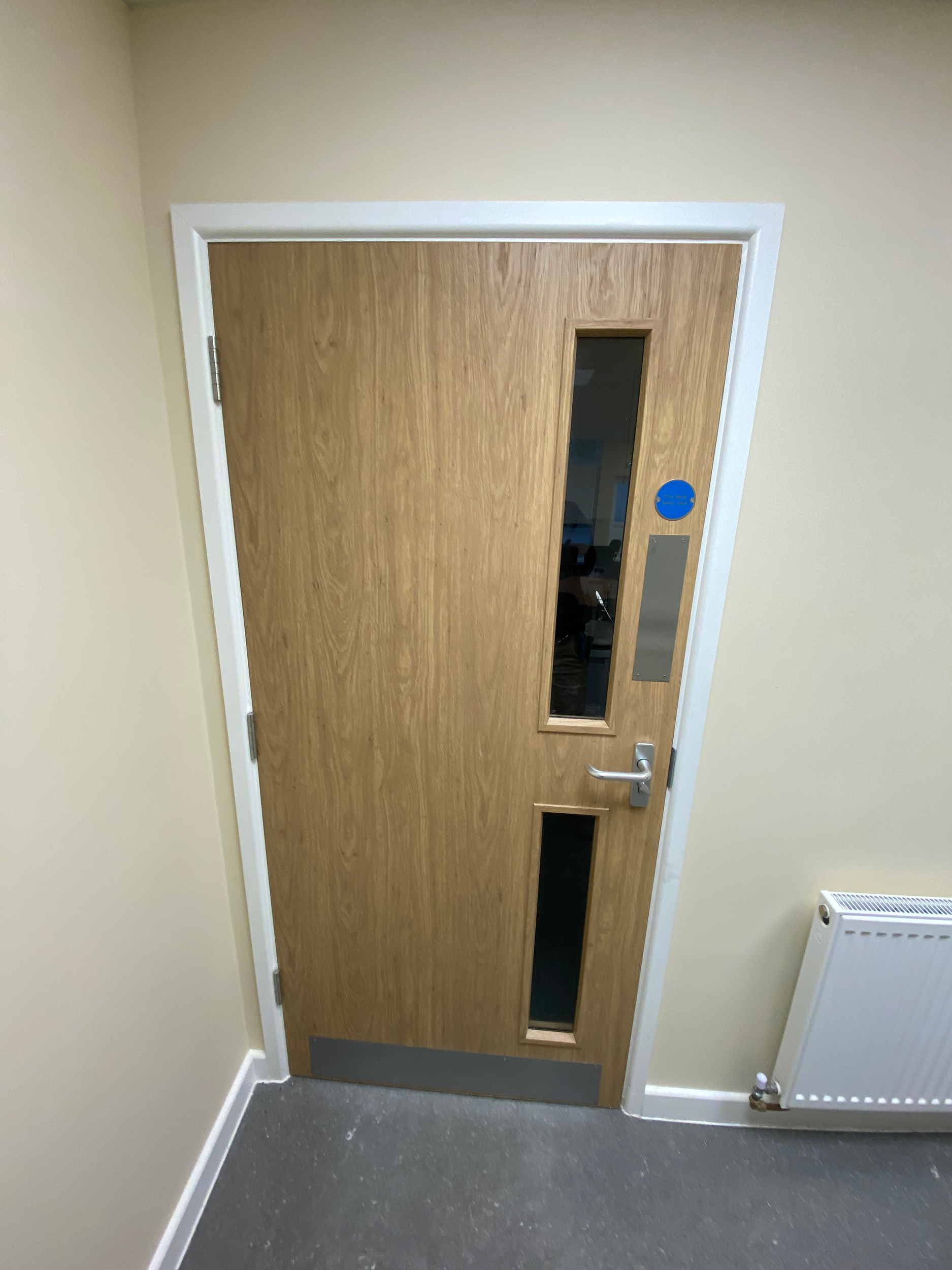 A wooden door with a vertical rectangular window near the top, a silver handle, and a grey kick plate at the bottom, set in a beige wall with white trim. To the right of the door is a white radiator.