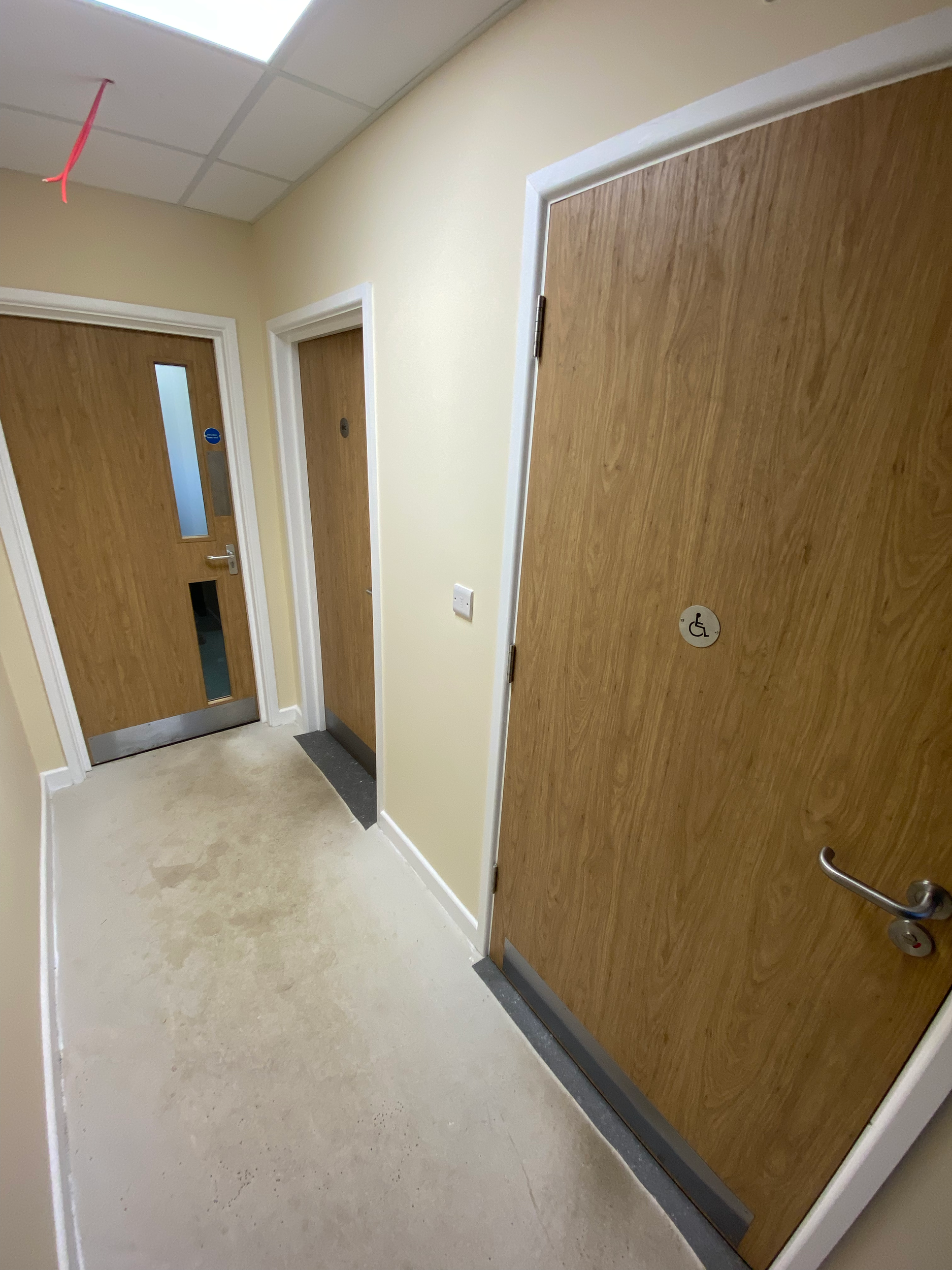Hospital hallway with wooden doors, one equipped with a wheelchair access sign, and a partially visible room number, beige walls, white trim, and a beige carpeted floor.