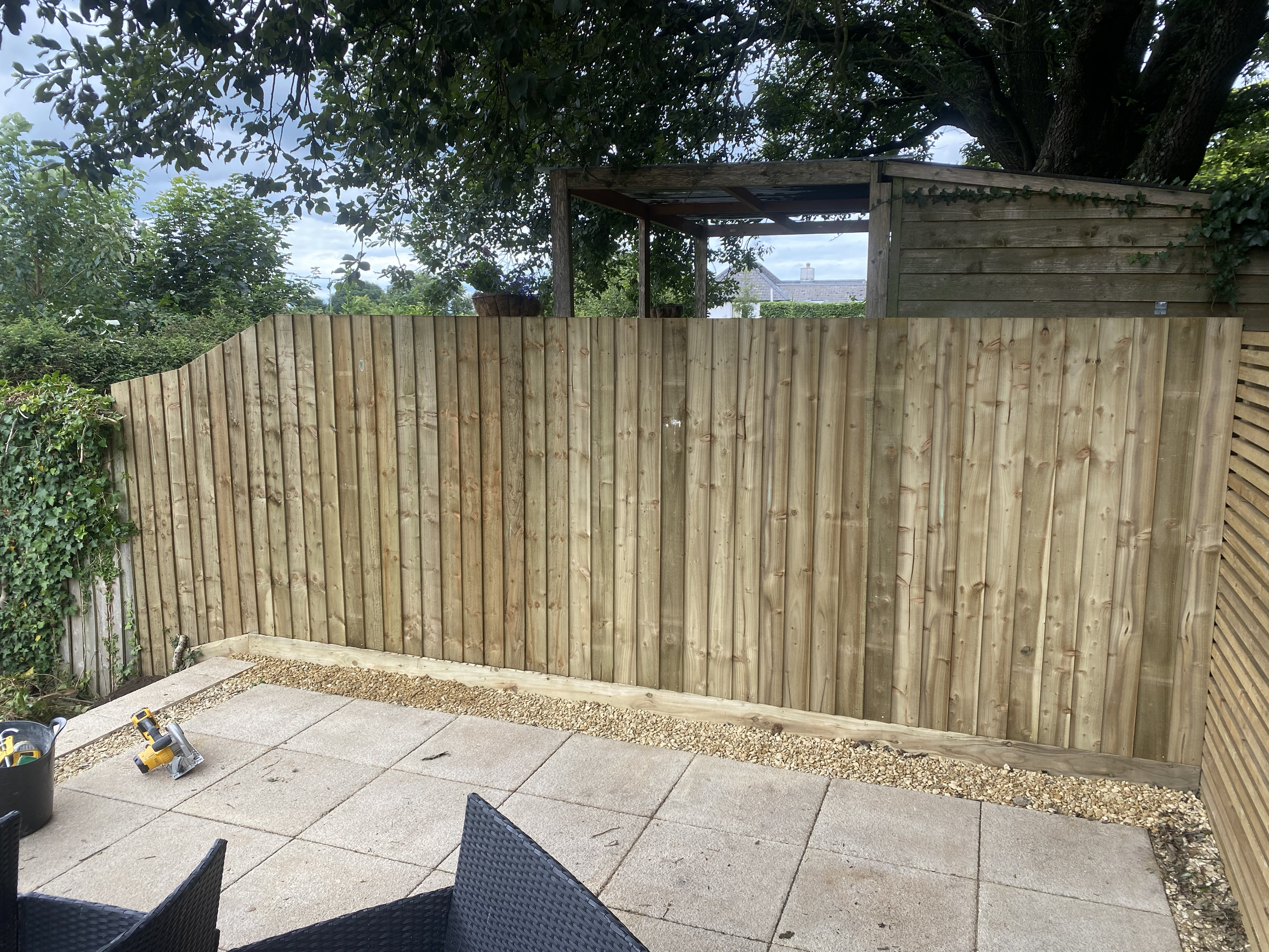 A wooden fence being built in a backyard with paving stones, construction tools, and a tree with leaves overhead.