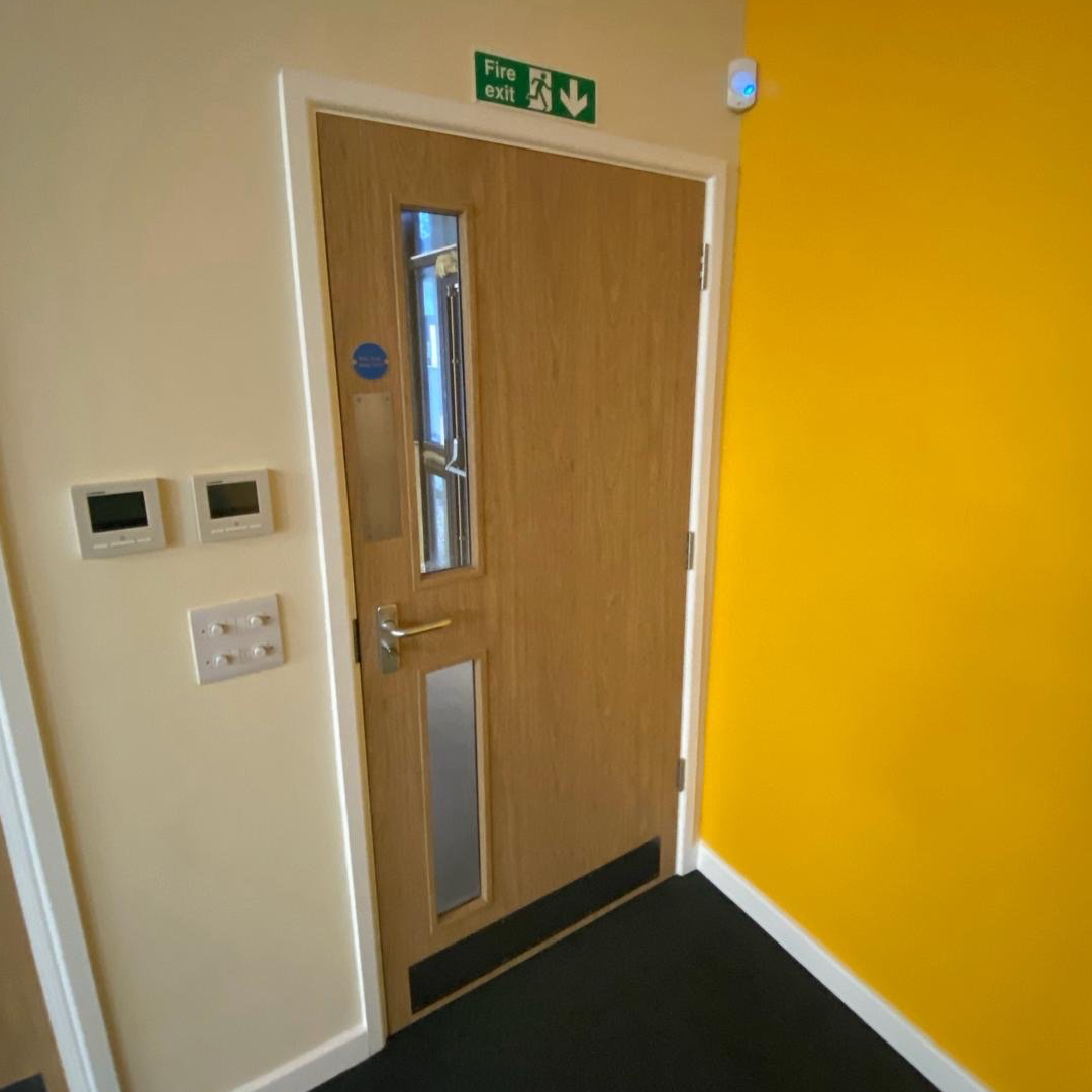Wooden door with a small window and a silver handle in a beige and yellow room, with a green fire exit sign above and a white security camera nearby.
