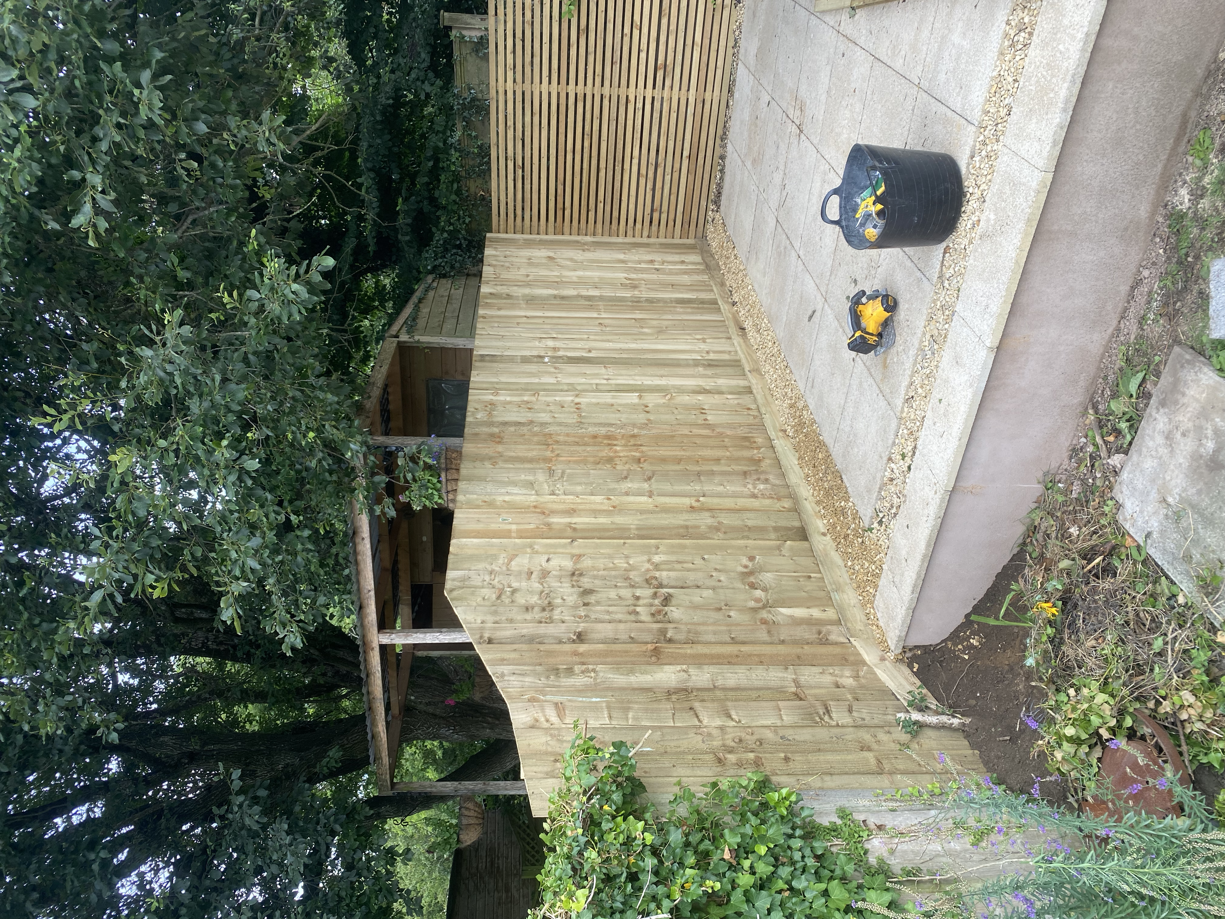 A backyard with a partially built wooden fence, a paved patio area, and gardening tools and supplies. Green trees and shrubs surround the space.