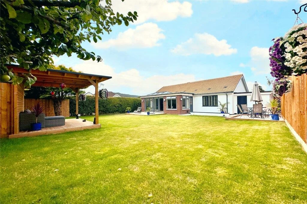 A spacious backyard with a well-maintained green lawn, a wooden gazebo on the left with outdoor seating, and a patio area on the right with a table and chairs under a large umbrella. The house has white walls, windows, and a tiled roof, with mountains visible in the distance under a partly cloudy blue sky.