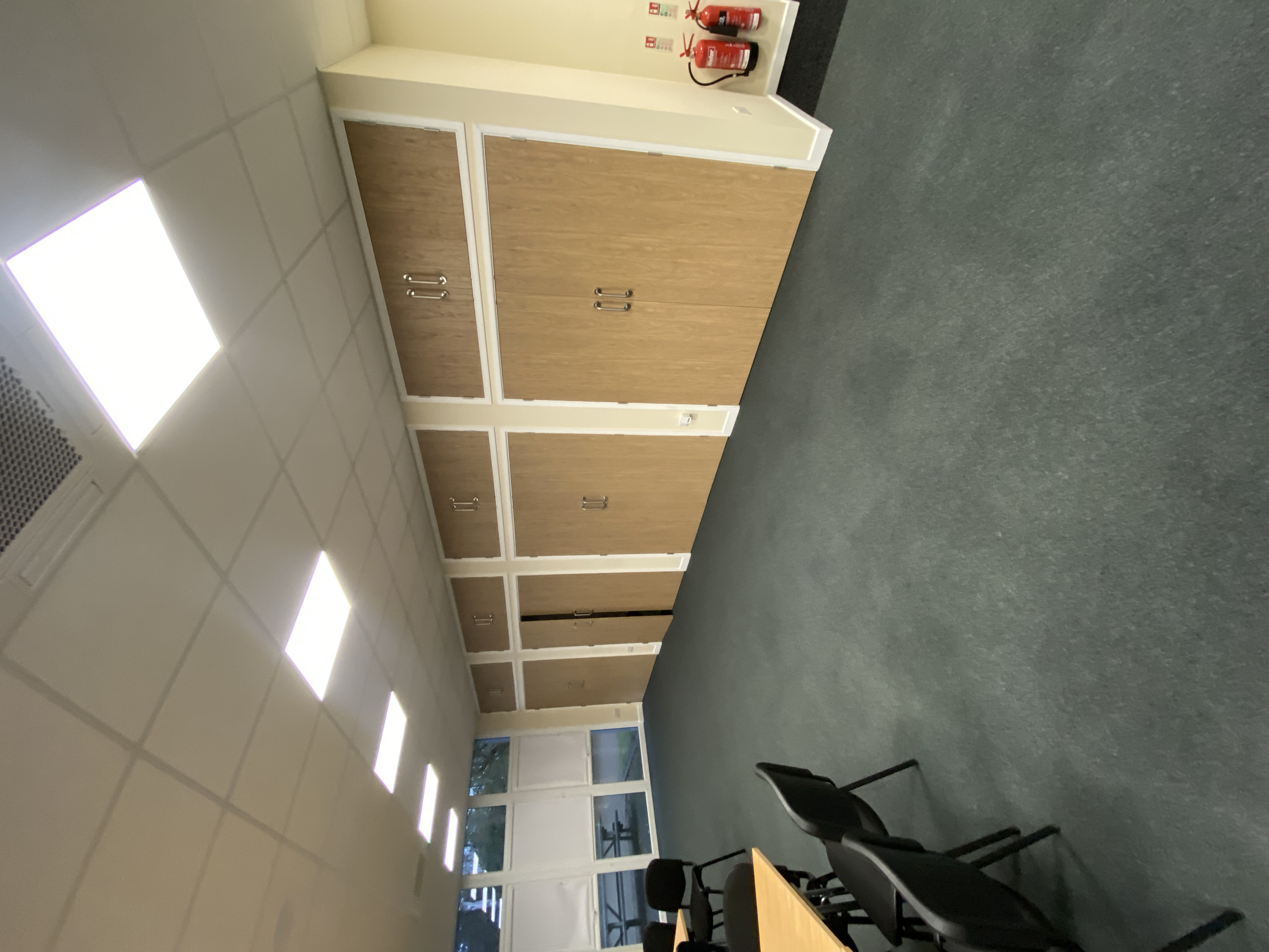Empty office room with wooden cabinets, black chairs, a table, and fire extinguishers on the wall. Fluorescent lighting on the ceiling and windows along one wall.