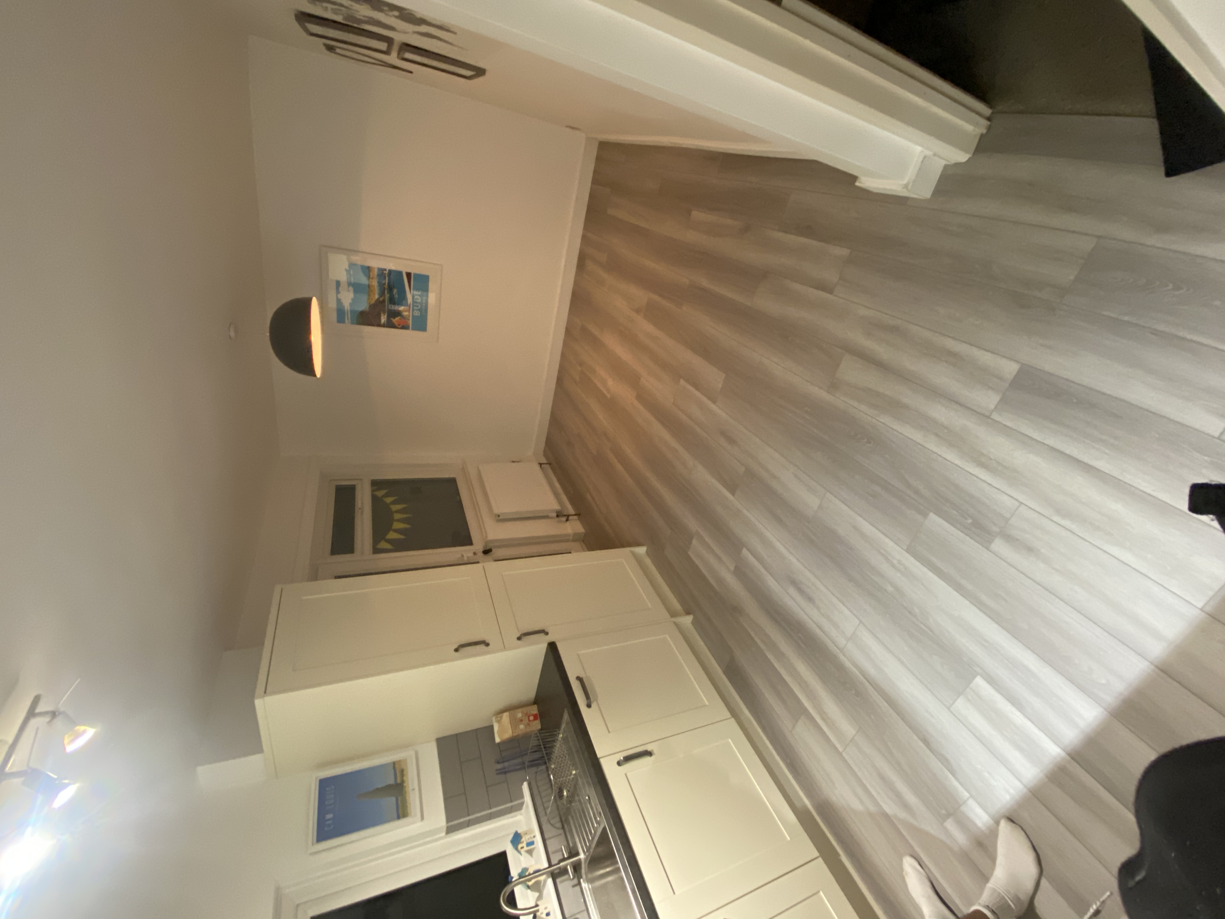 A kitchen with white cabinets, gray countertops, a window with a sun design on the blinds, and light wood flooring. A poster of a lighthouse on the wall, a hanging black pendant light, and a person wearing white socks are visible.