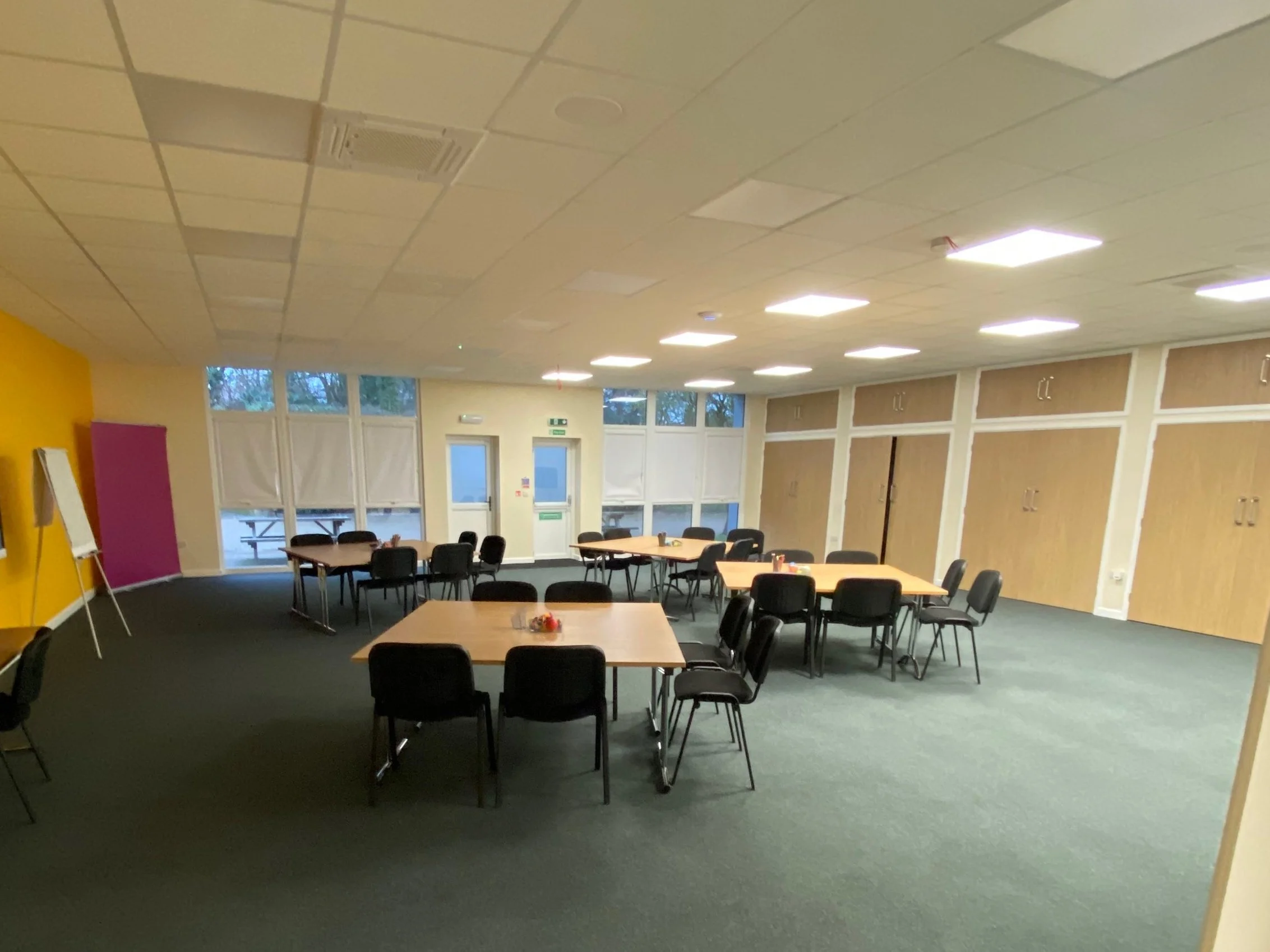 A spacious conference room with several tables and chairs arranged for a meeting or event. The room has a yellow and purple accent wall, large windows with blinds, and wooden cabinets along the wall. There are ceiling lights and an easel with a flip chart stand.