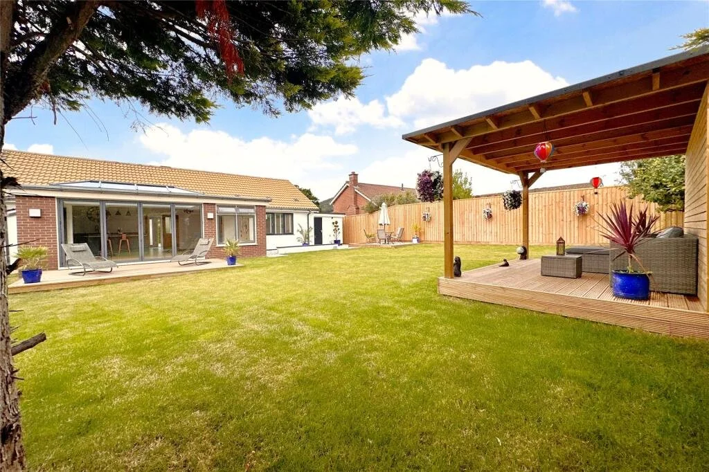 A backyard with a well-maintained green lawn, a brick house with a large glass sliding door, an outdoor seating area on a wooden patio with a covered roof, potted plants, hanging flower baskets, and a wooden fence.