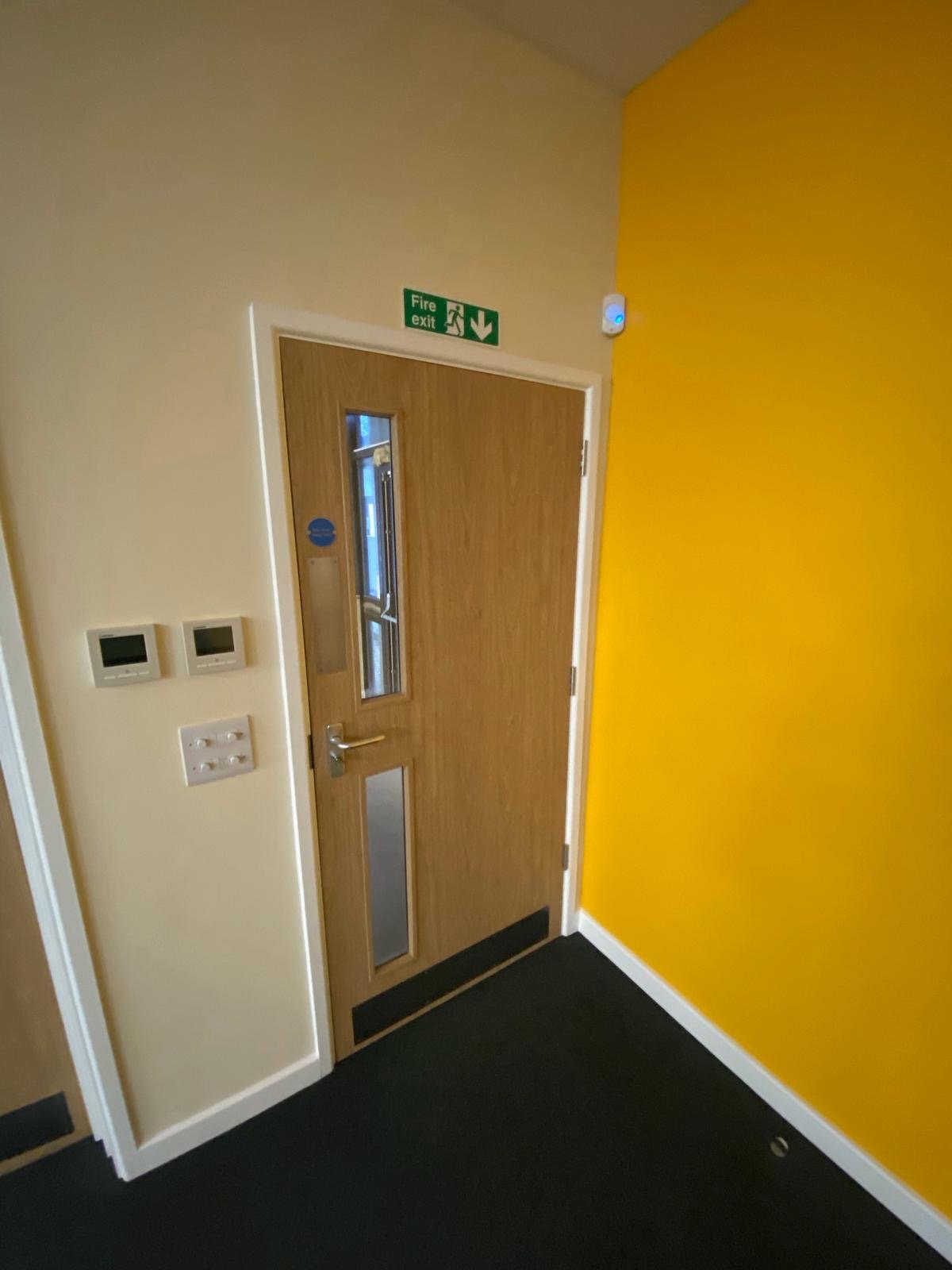 Wooden door with window, yellow wall on the right, white wall on the left, green fire exit sign above door, black carpeted floor.