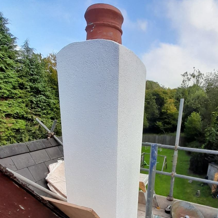 Chimney with freshly painted white base and unpainted top on a rooftop with scaffolding, roof shingles, and green trees in the background.