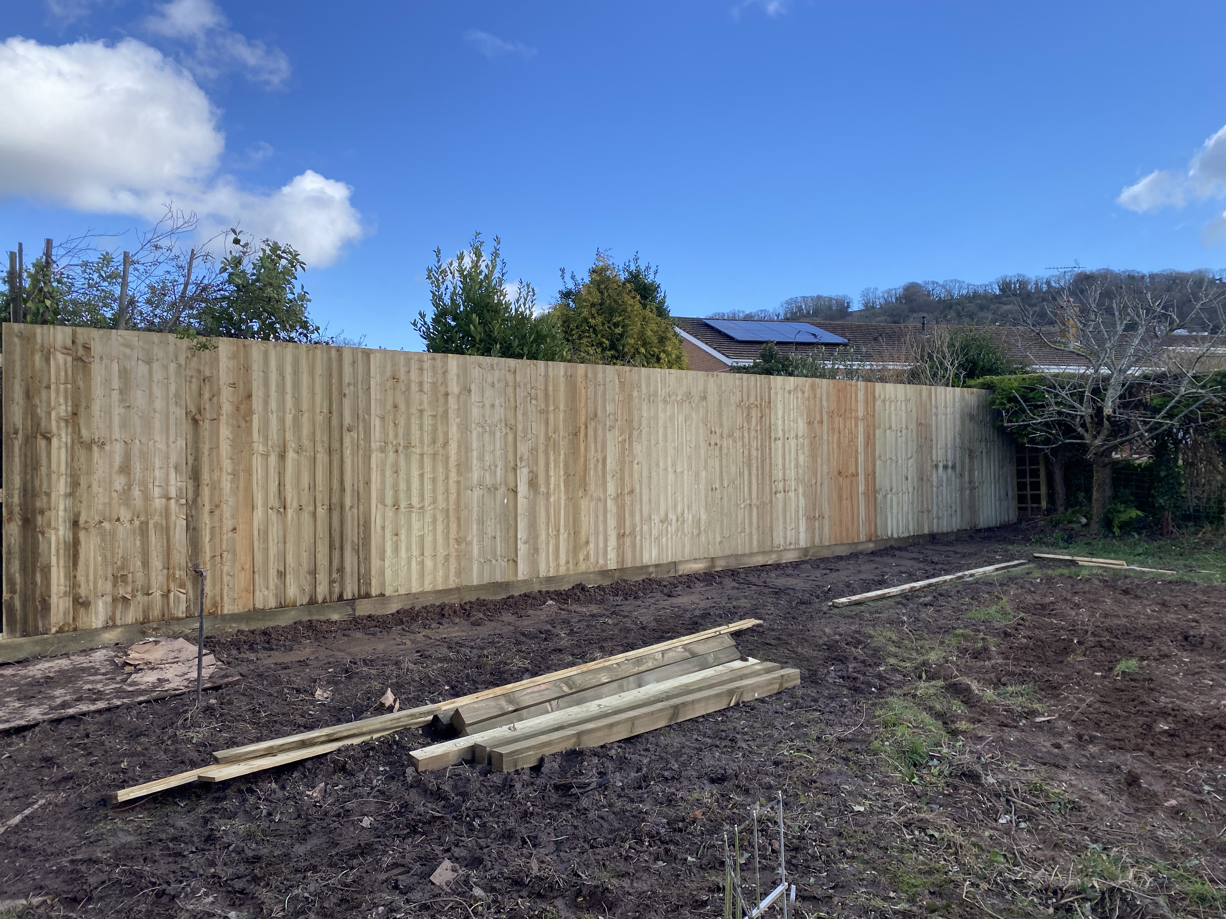 A new wooden fence installed in a backyard, with some construction materials and wood planks on the ground in front, under a partly cloudy blue sky.