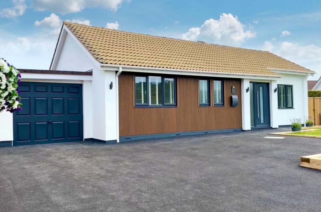 A modern house with a brown tiled roof, white walls, and a dark blue garage door. The house has multiple windows with blinds and a front door with a glass panel. The driveway is paved, and there are some plants and a flower basket hanging on the side
