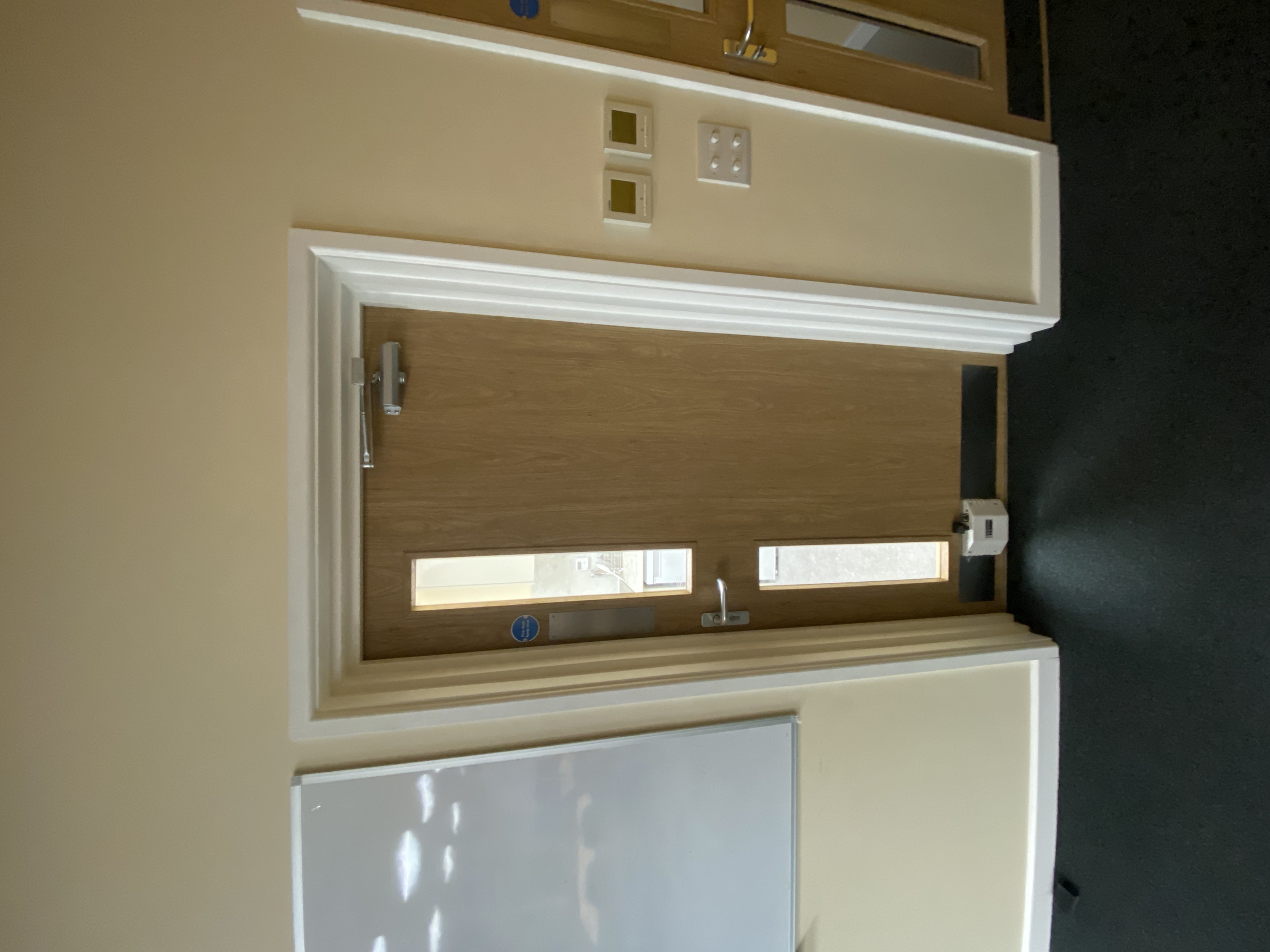 Wooden door with a vertical window, door handle, and a push bar, set in a beige wall with electrical switches and a fire alarm, adjacent to a whiteboard and a dark-colored floor.