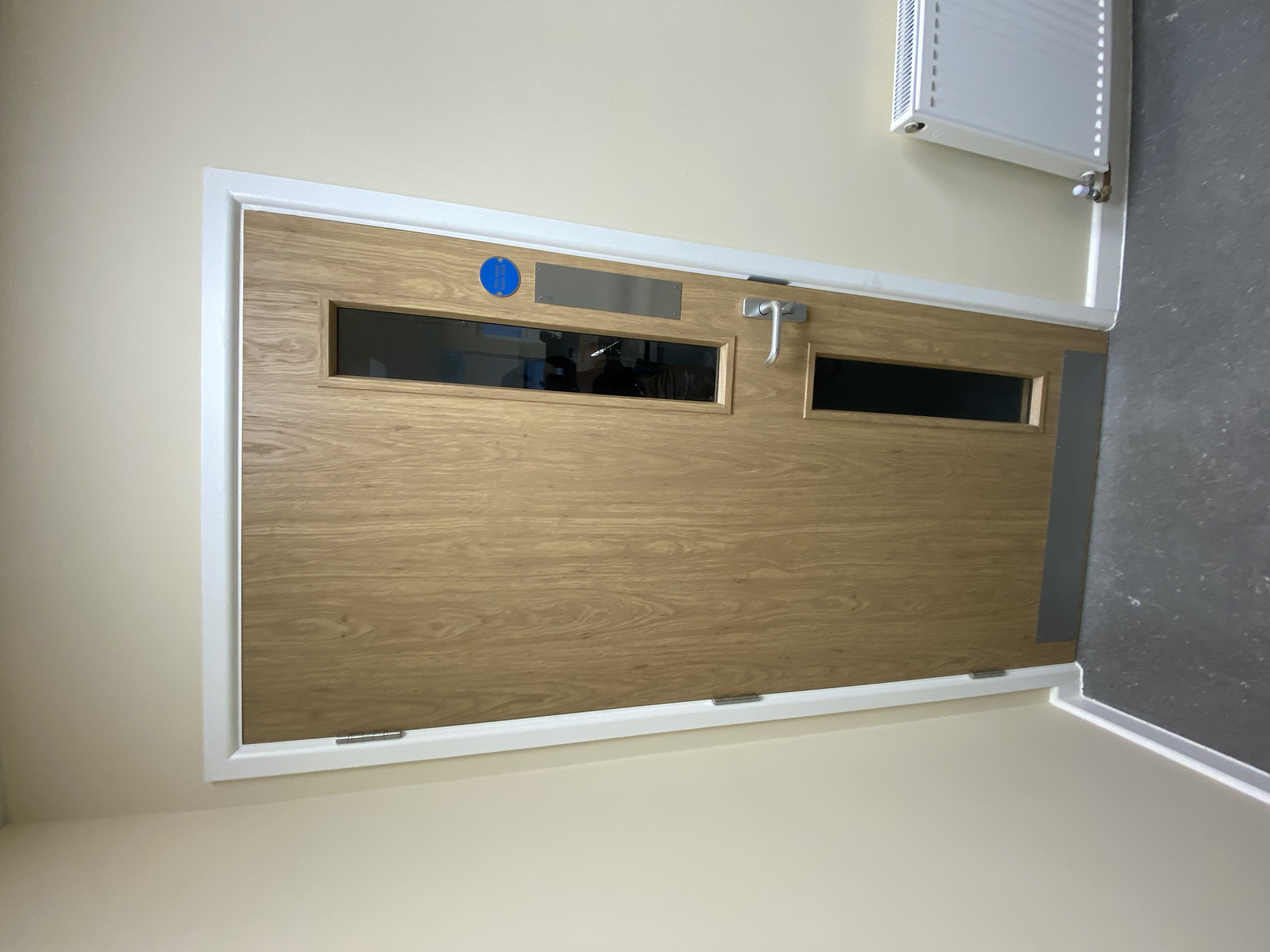 A wooden door with a vertical rectangular window near the top, a silver handle, and a grey kick plate at the bottom, set in a beige wall with white trim. To the right of the door is a white radiator.