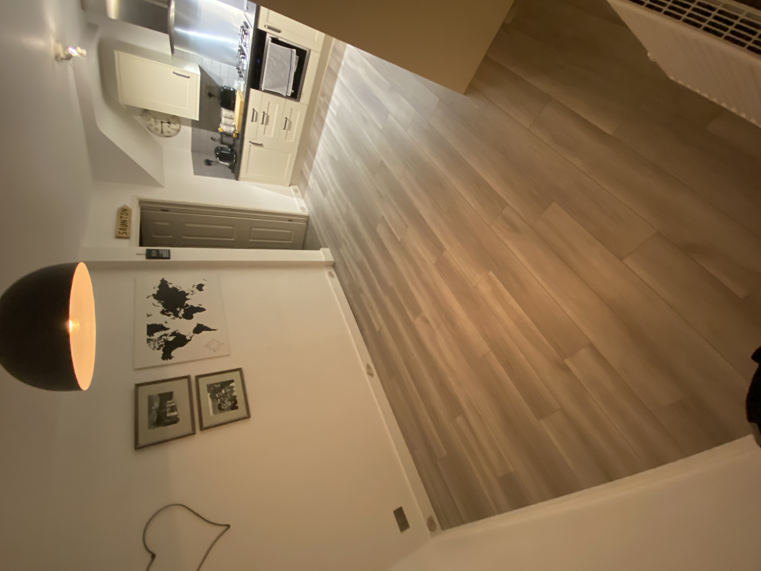 View of a kitchen with white cabinets, a stove, a sink, and countertops. The room features wooden flooring and wall decor including a world map and framed photos. Part of a dark hanging lamp and an abstract wall outline can be seen.