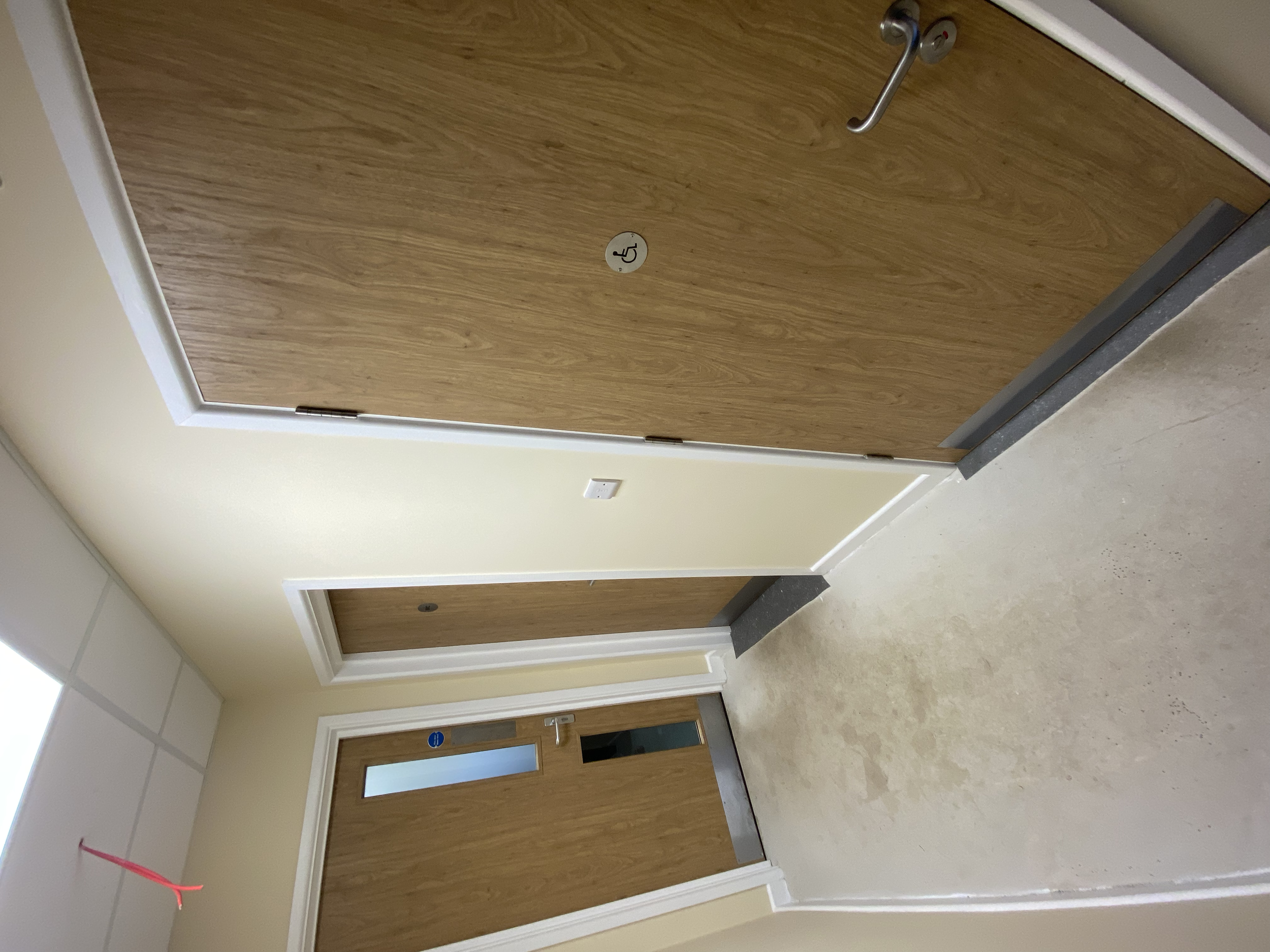 Hospital hallway with wooden doors, one equipped with a wheelchair access sign, and a partially visible room number, beige walls, white trim, and a beige carpeted floor.