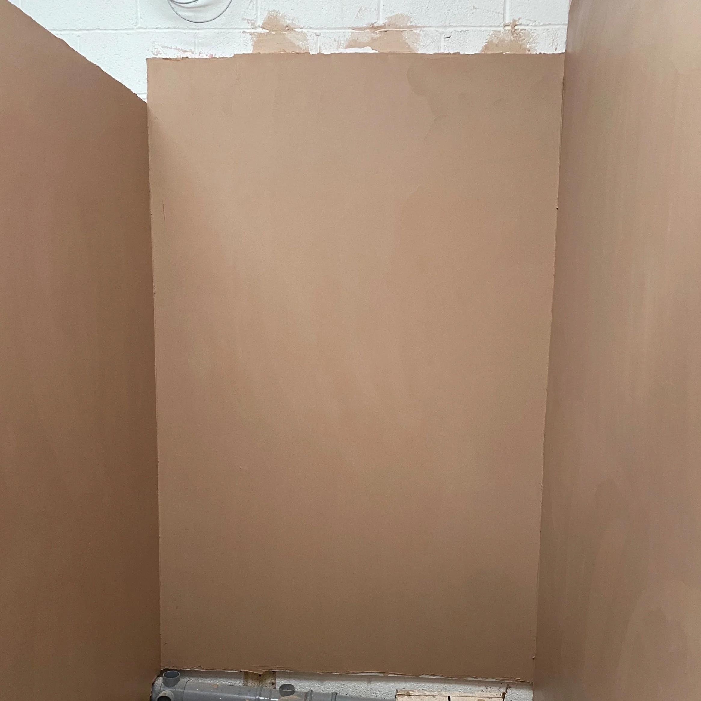Interior view of a partially constructed room with brown drywall panels installed on the walls and an unfinished white brick ceiling. There is a small section of flooring visible with construction materials at the bottom.