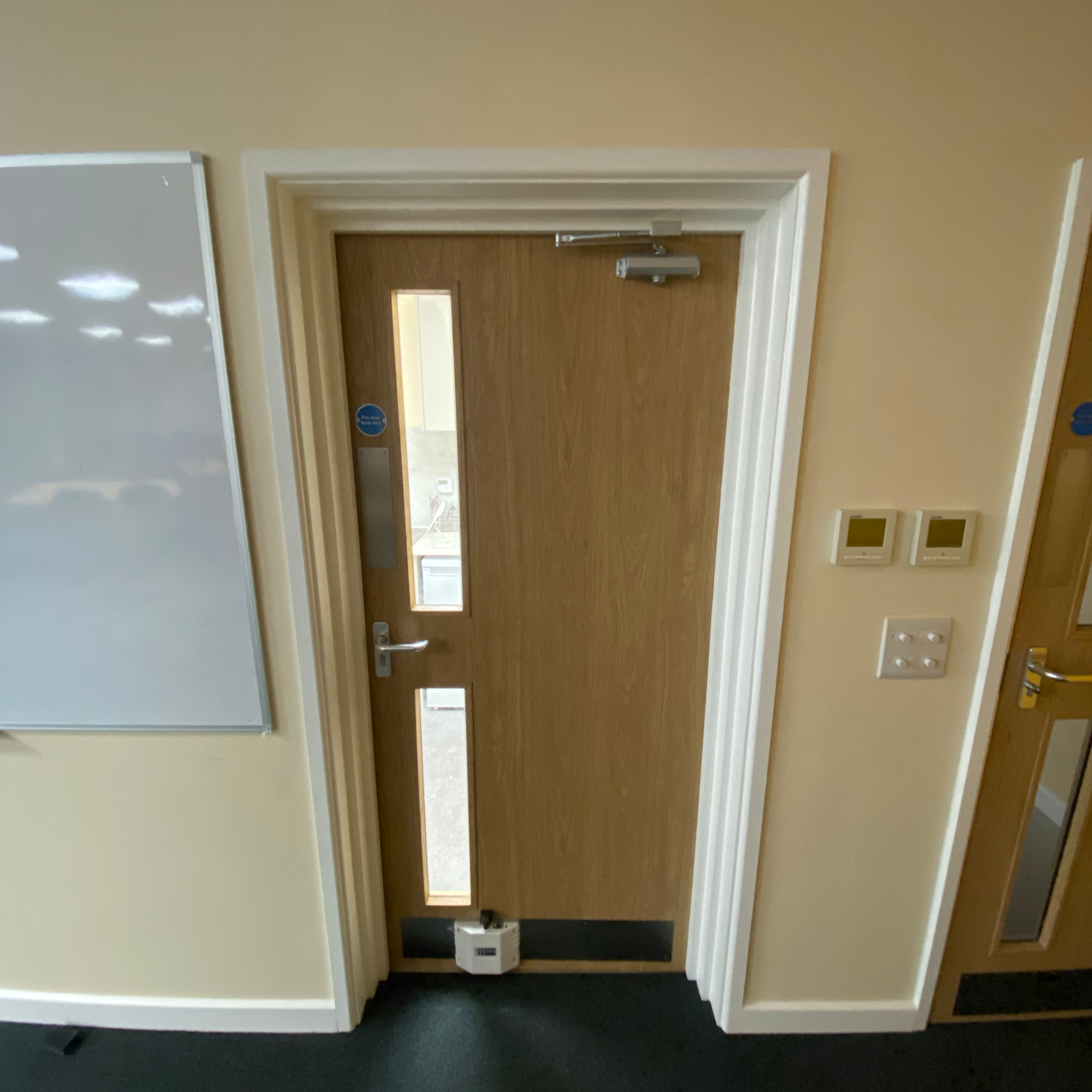 Interior view of a wooden door with two vertical rectangular windows, white doorframe, and security device at the top, set in a light-colored wall with two thermostat controls and a light switch nearby.