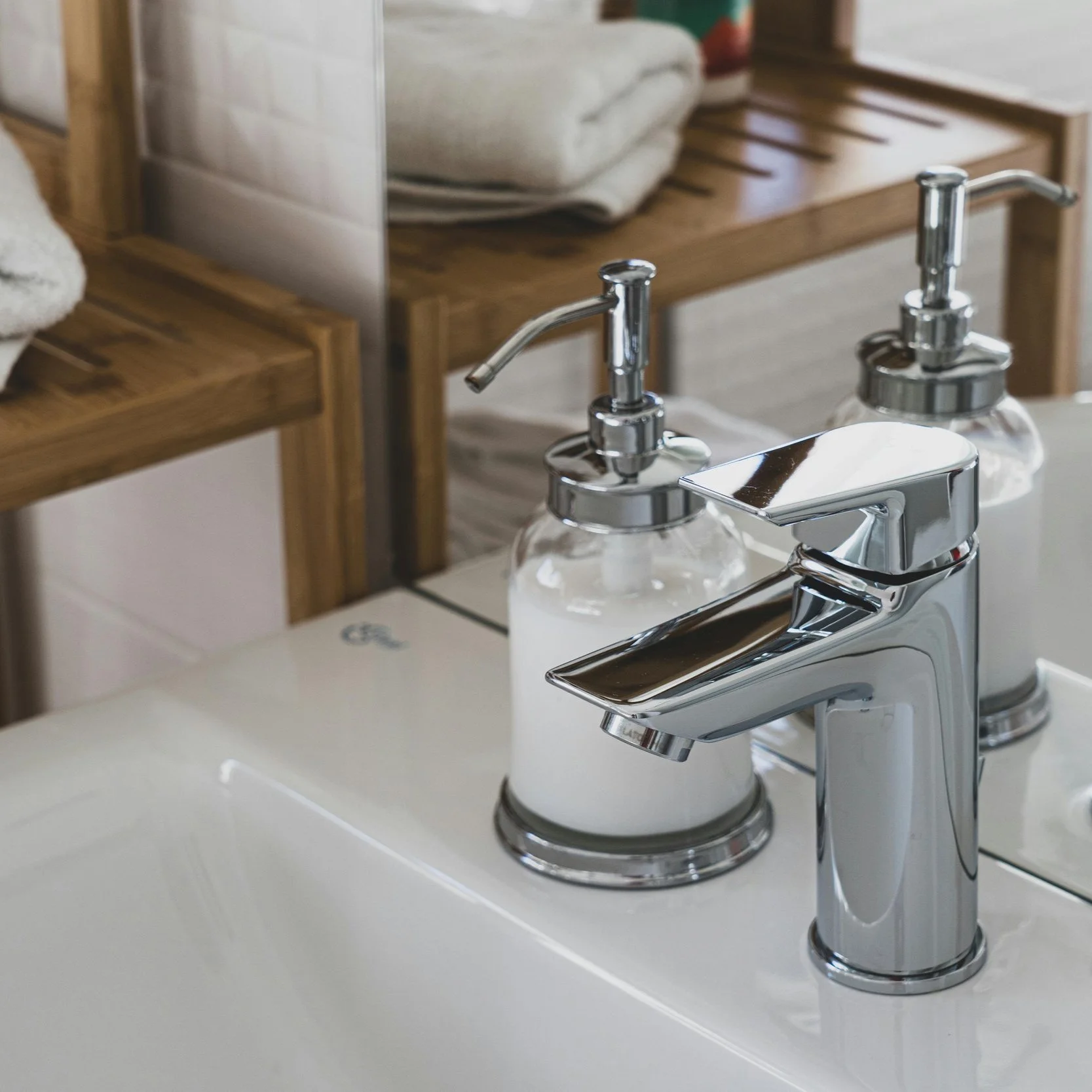 Close-up of a modern chrome bathroom sink faucet with two glass soap dispensers behind it, on a white sink with bathroom accessories and wooden furniture in the background.