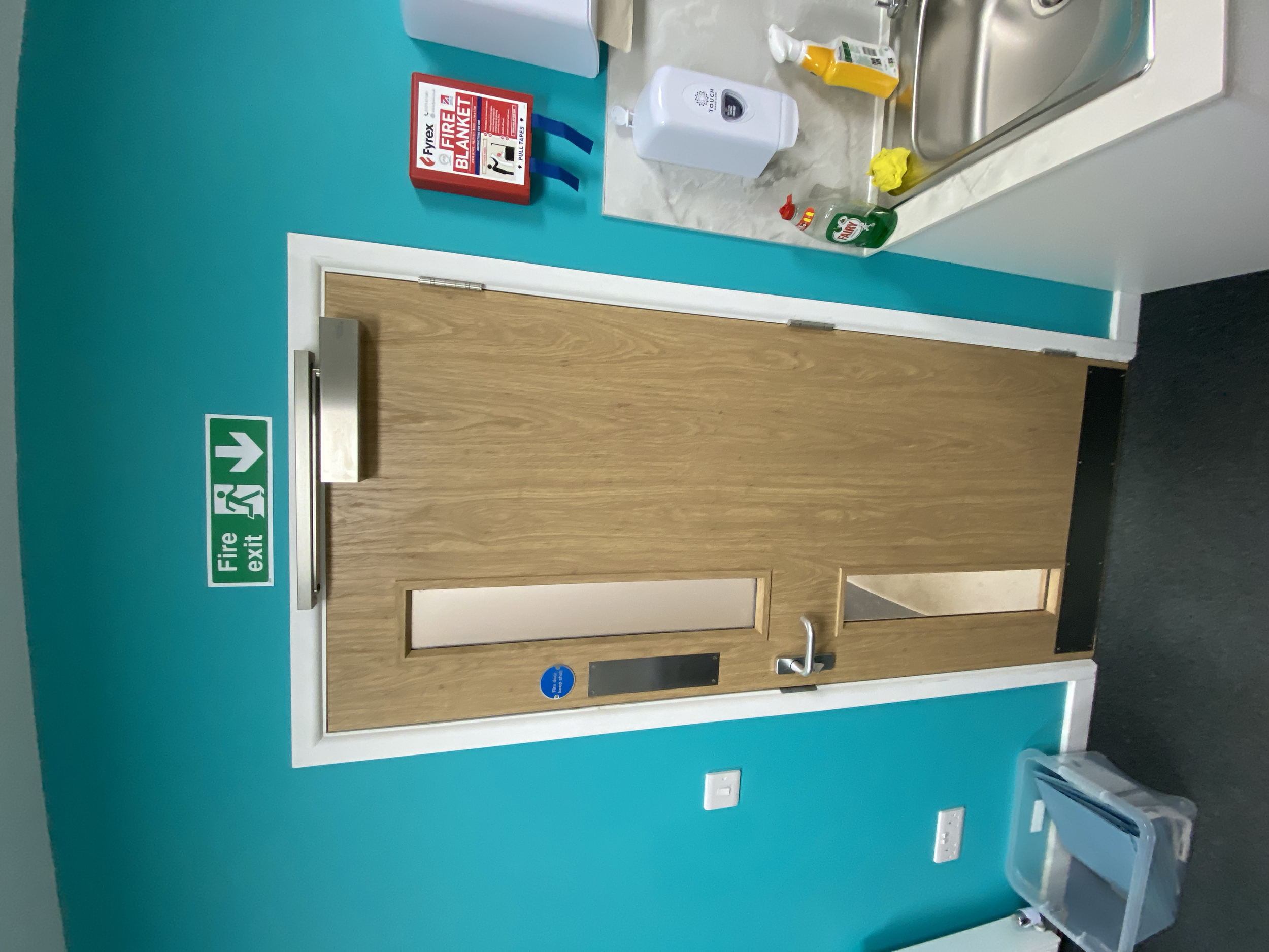 Modern kitchen with blue walls, a wooden door with windows, a stainless steel sink, a fire extinguisher box, cleaning supplies on the counter, and a plastic storage container on the floor.